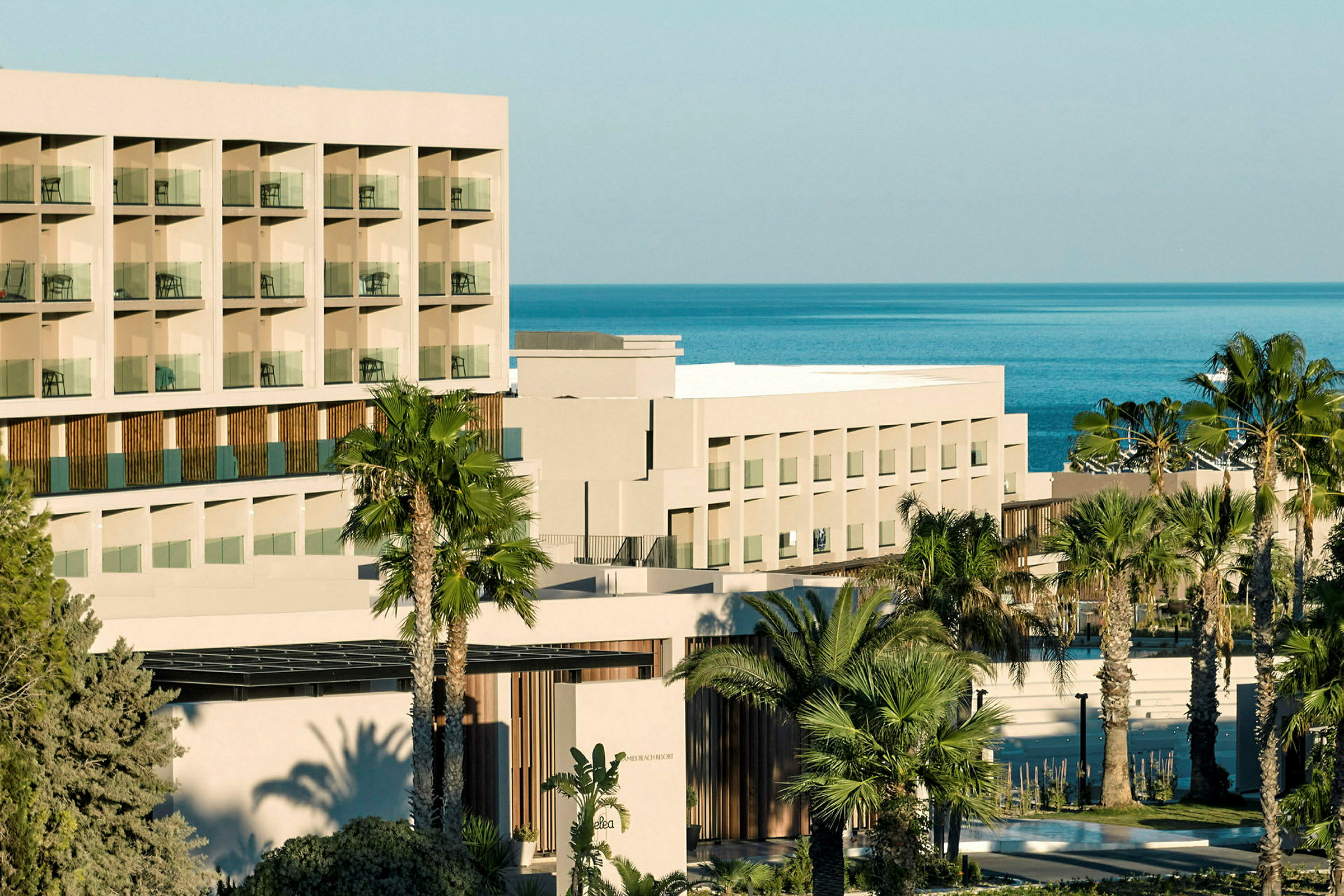 Bilde av en moderne hotellbygning med flere etasjer og balkonger, omgitt av palmer og med utsikt over havet. (AI-generert tekst)