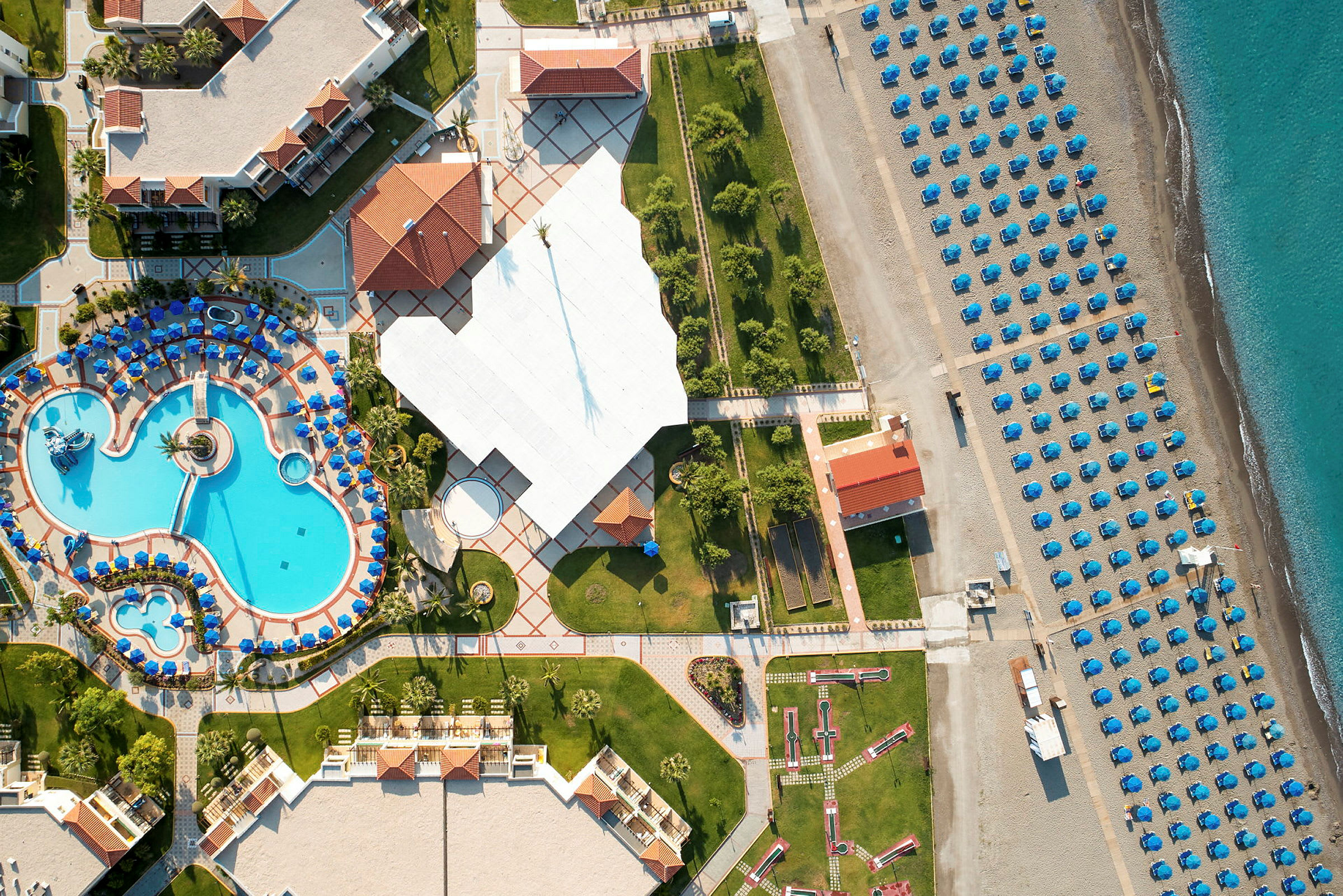 Flyfoto som viser et feriehotell med bassengområde, grøntområder og en strand med oppstilte parasoller. (AI-generert tekst)
