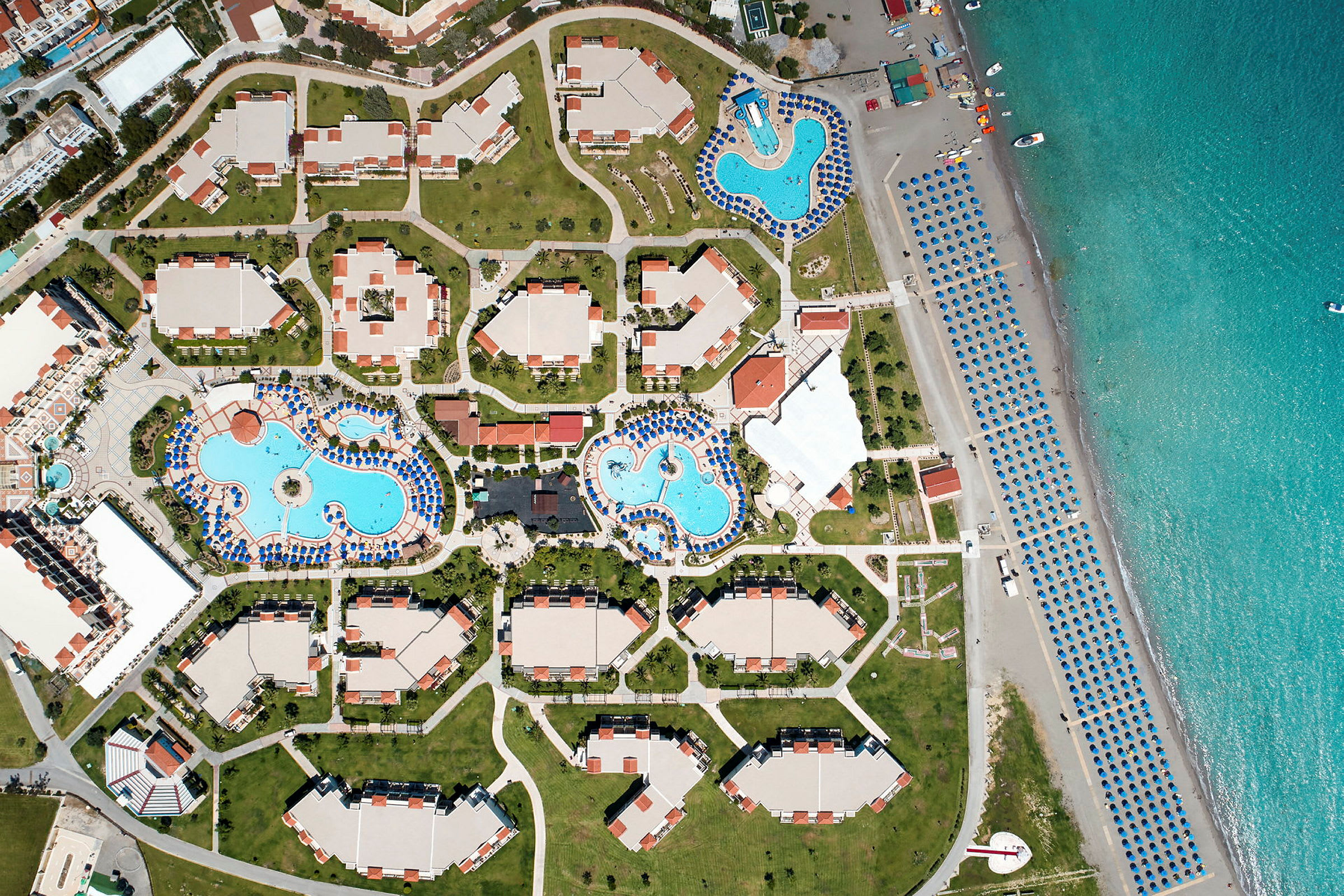Fugleperspektiv av en resort med flere bygninger, bassengområder og strand med parasoller. (AI-generert tekst)
