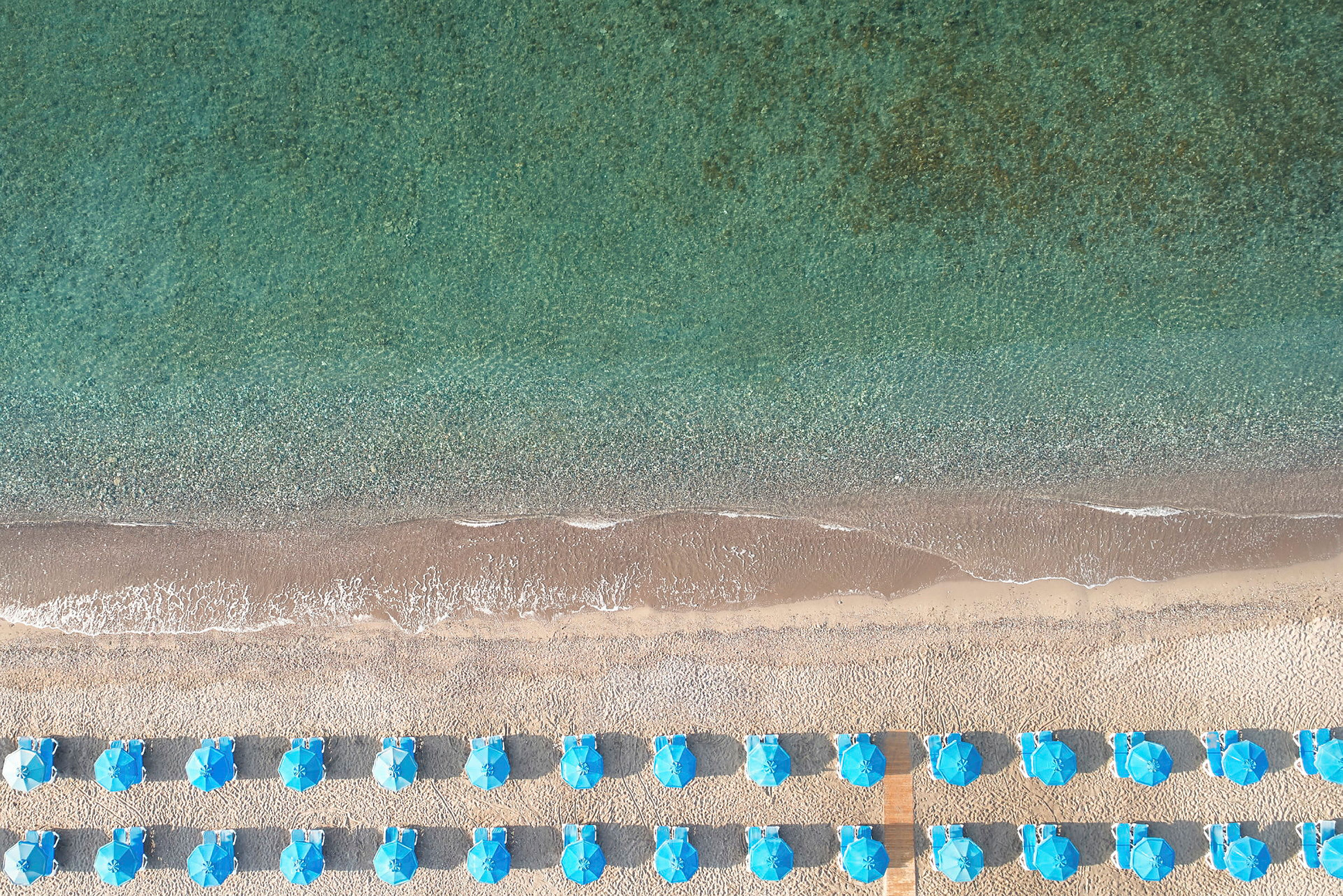 Flyfoto av en strand med blå parasoller og turkist hav. (AI-generert tekst)