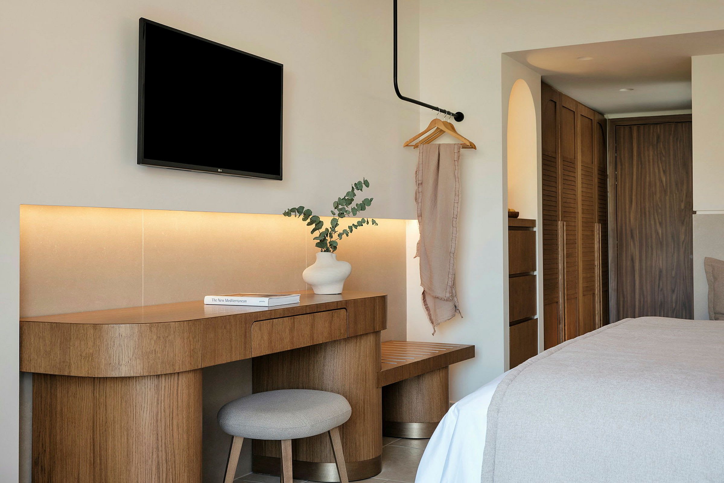 Et hotellrom med moderne interiør. Et trebord med en hvit vase og en bok står ved siden av en stol. En svart TV er montert på veggen over. (AI-generert tekst)