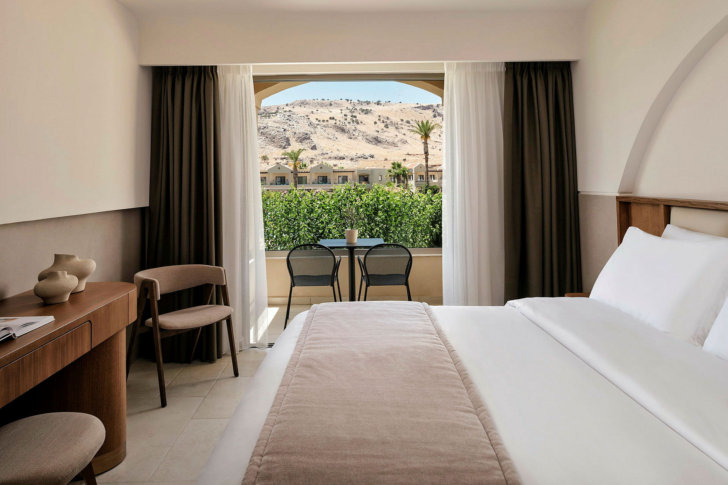 Hotellrom med en stor seng, skrivebord og stol. Utsikt mot grønne områder og fjell gjennom glassdører. (AI-generert tekst)