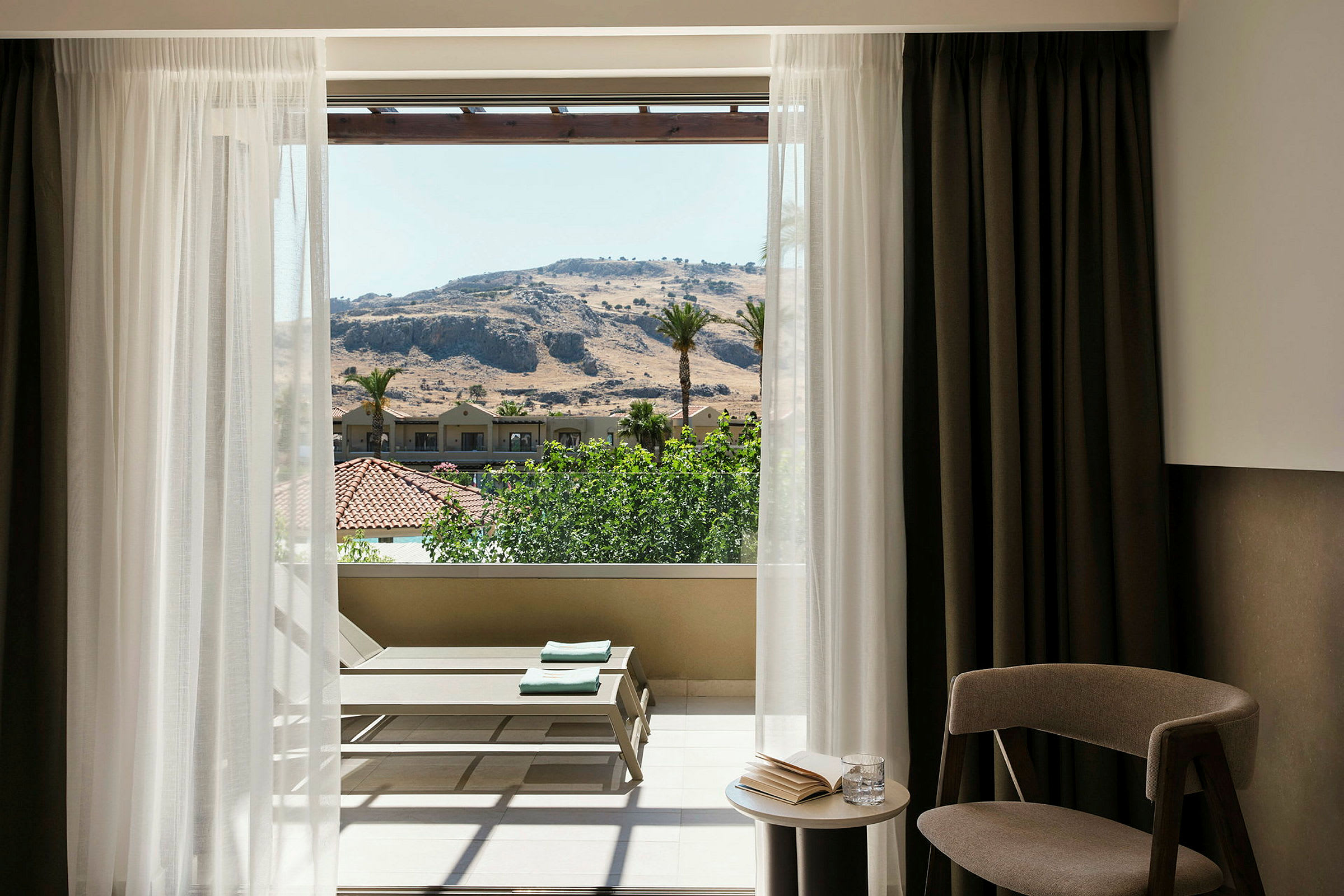 Et hotellrommiljø med en åpen balkongdør som viser utsikten over et fjellandskap og grøntområder. En lenestol og et lite bord er plassert nær balkongen. (AI-generert tekst)