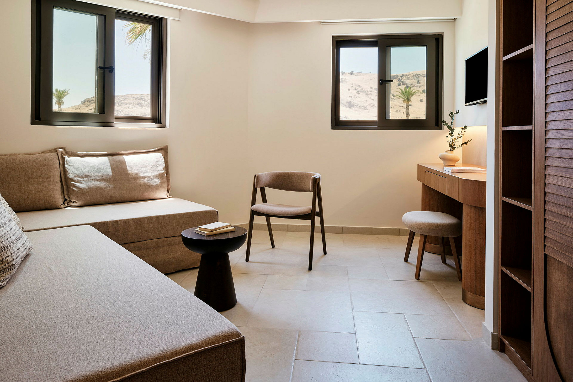 Et hotellrom med sofa, stol, bord og skrivebord, samt vinduer med utsikt over et tørt landskap. (AI-generert tekst)