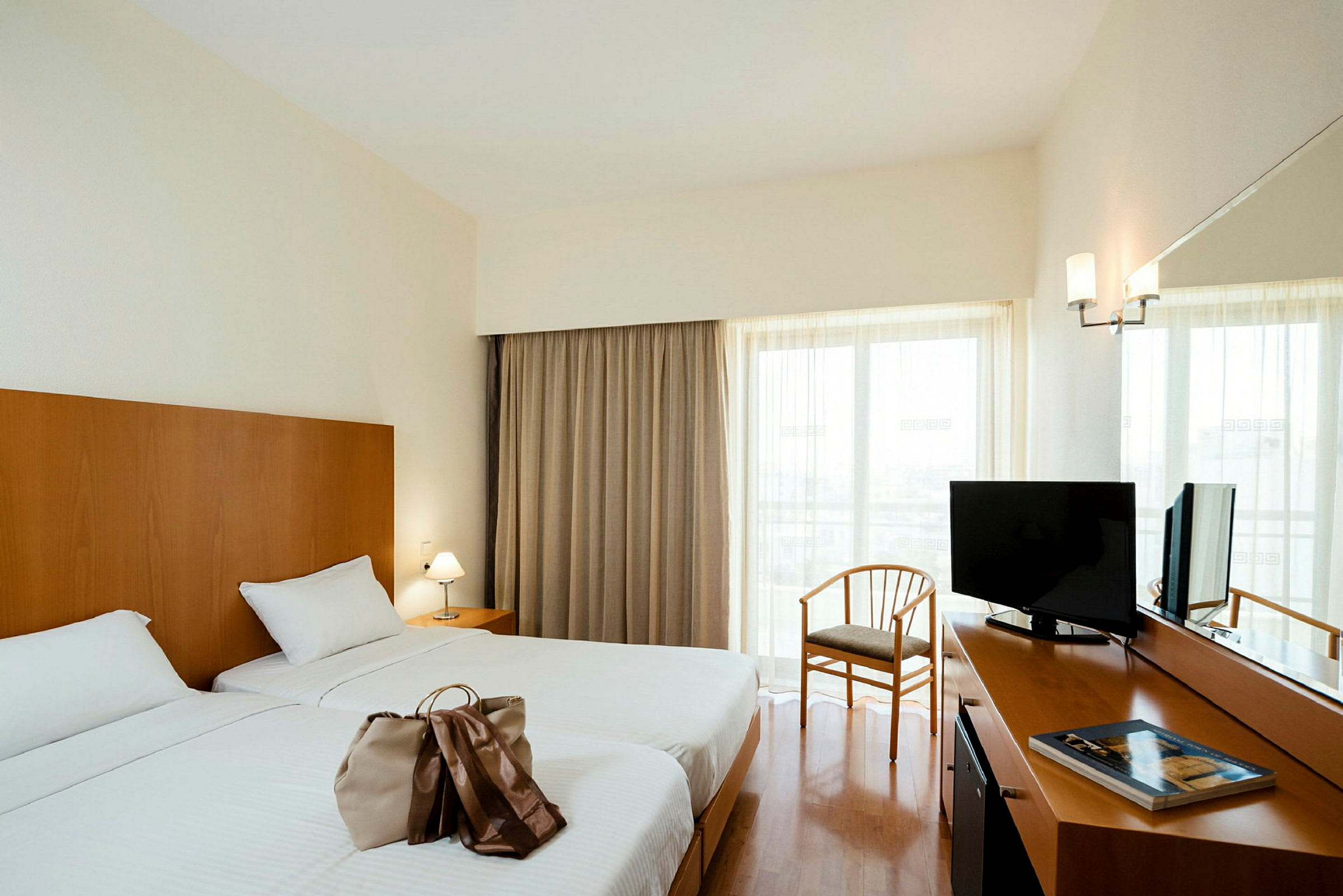 Hotellrom med to enkeltsenger, et skrivebord med TV og en stol foran et vindu med gardiner. (AI-generert tekst)
