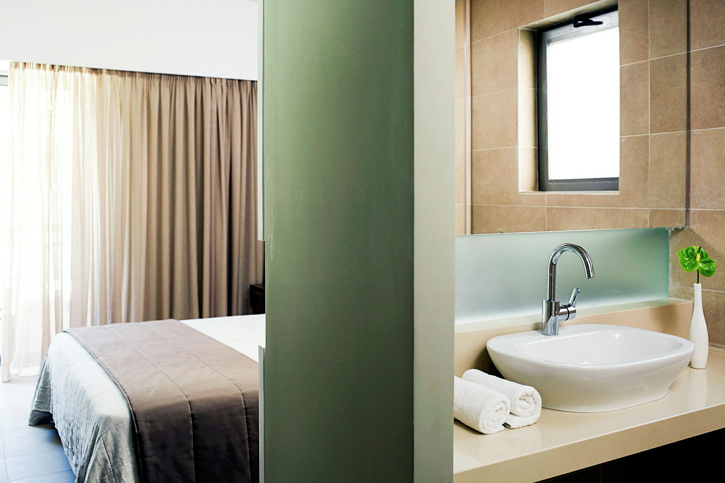 Et hotellrom med en delt visning mellom soverom og bad. Soverommet har en seng med beige sengetøy og et vindu med gardiner. Badet har en hvit vask, håndklær og et lite grønt blad i en vase. (AI-generert tekst)