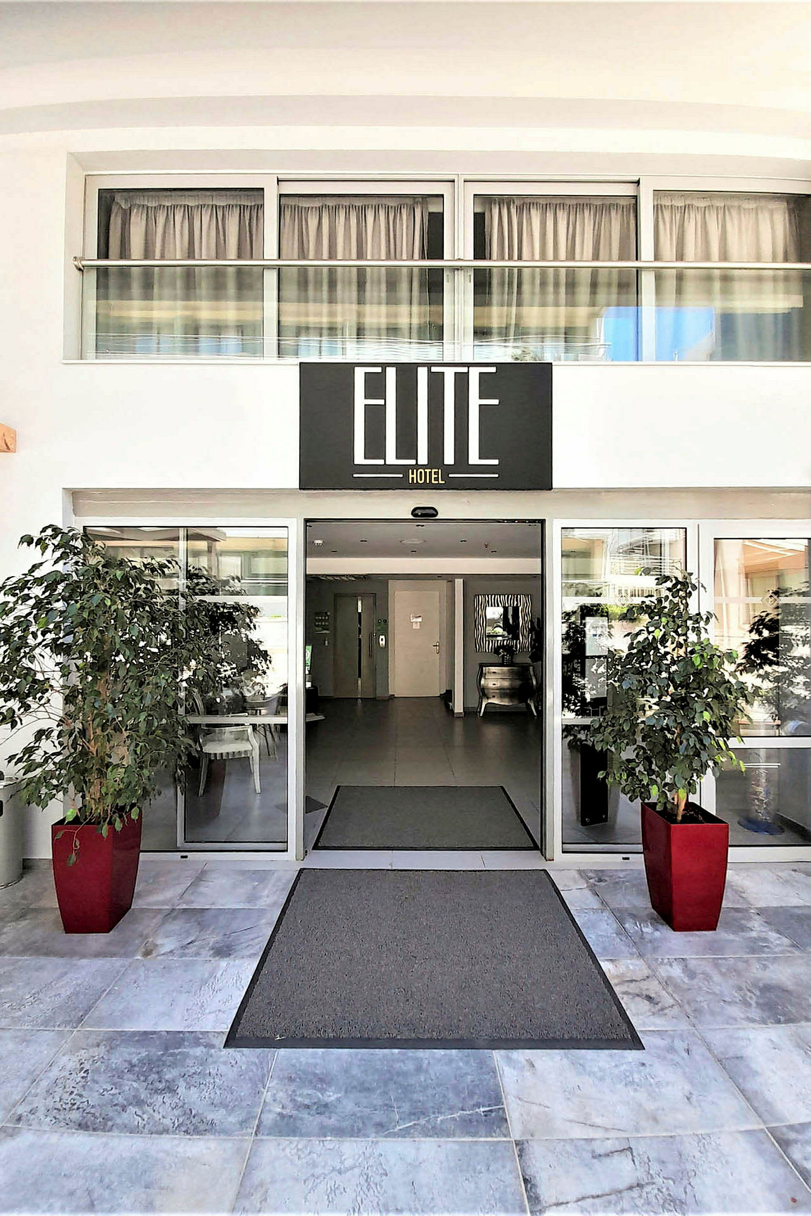 Inngangen til Elite Hotel med glassdører og røde blomsterpotter. (AI-generert tekst)