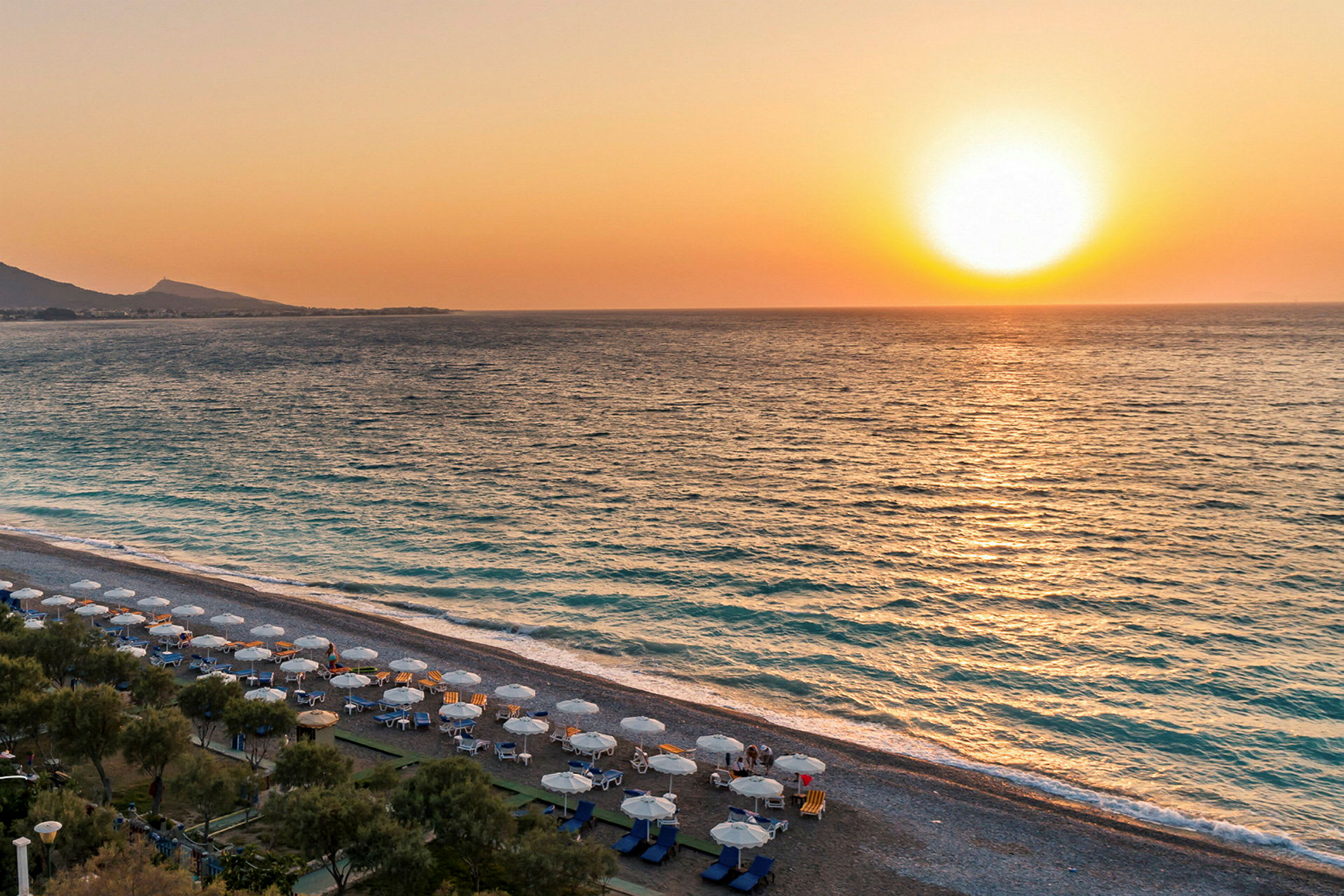 Solnedgang over havet med en strand i forgrunnen utstyrt med solsenger og parasoller. (AI-generert tekst)