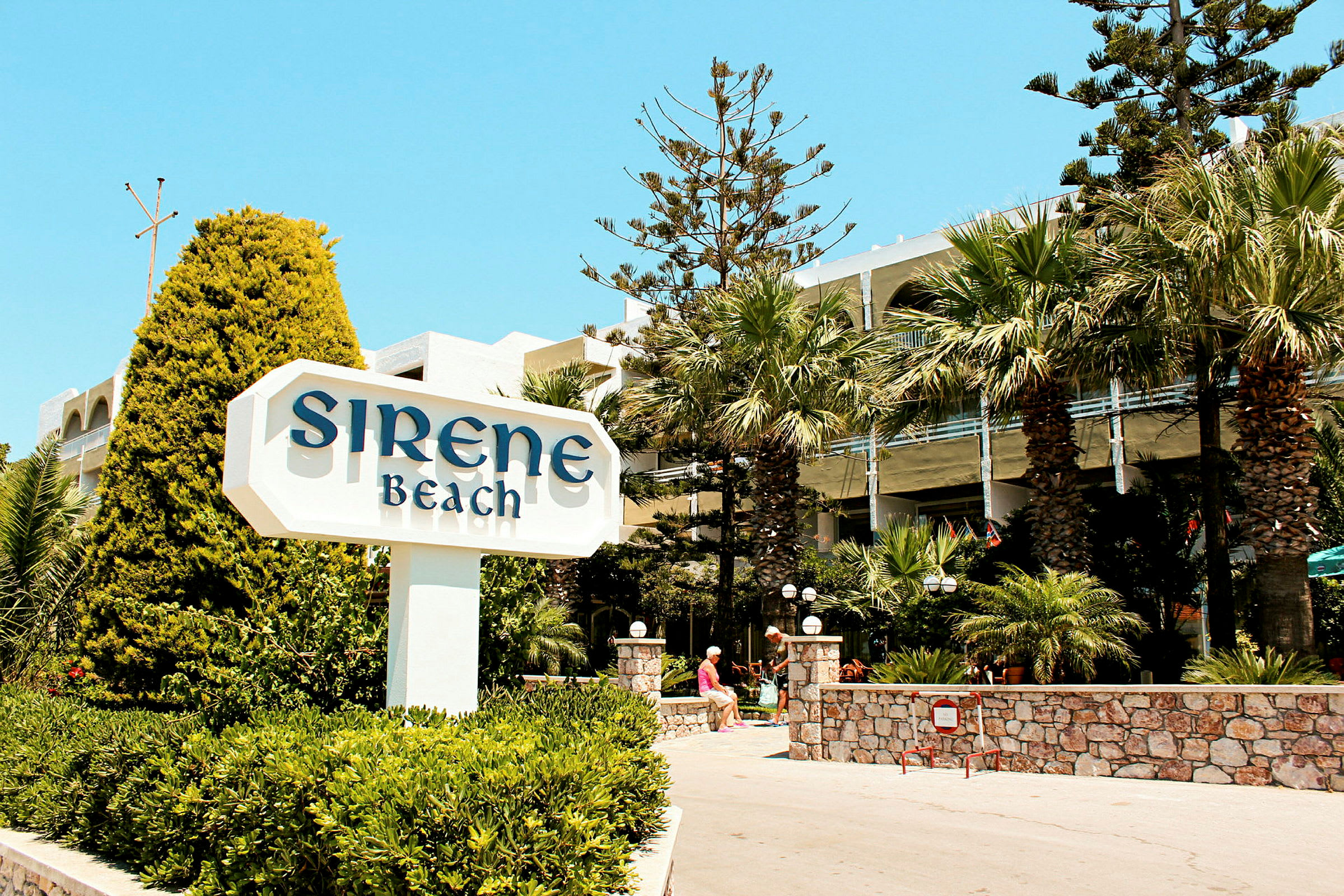 Skilt med teksten 'Sirene Beach' foran et hotell med palmer og vegetation. (AI-generert tekst)