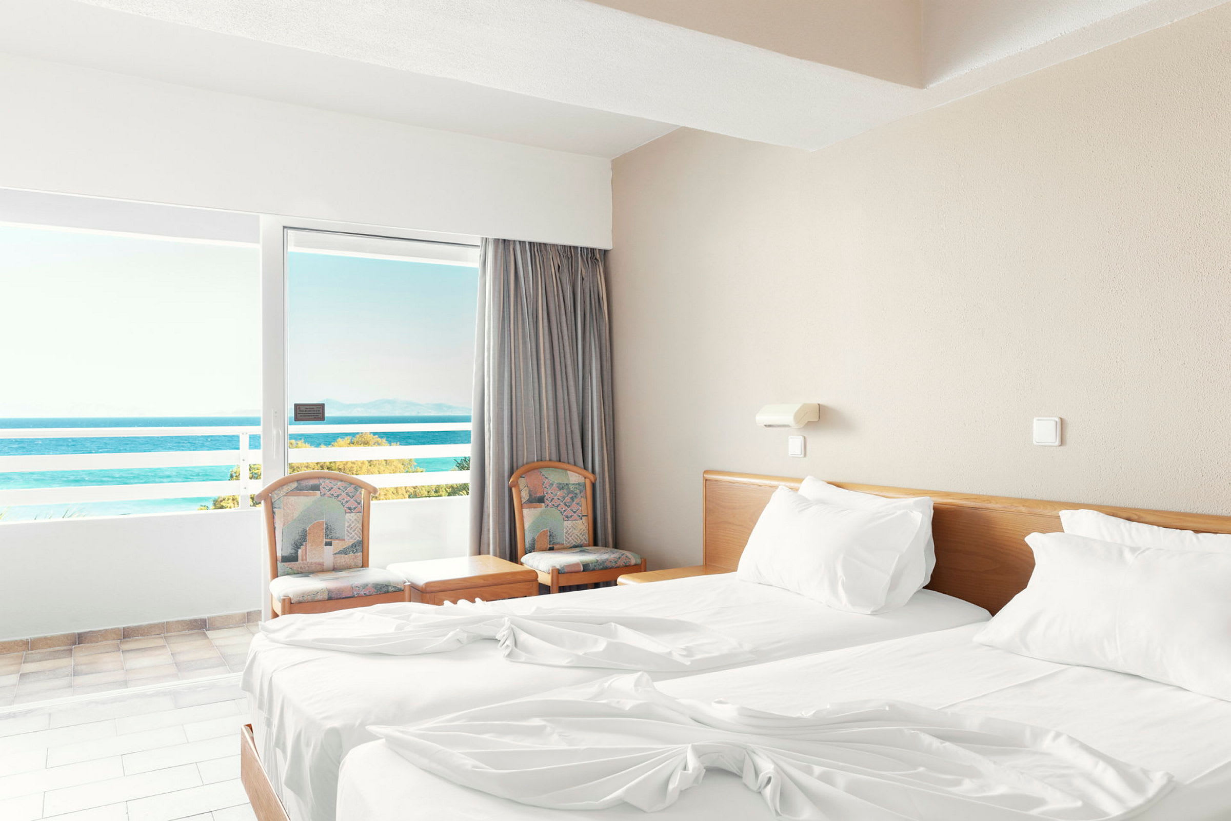Et hotellrom med to enkeltsenger og en balkong med utsikt over havet. (AI-generert tekst)