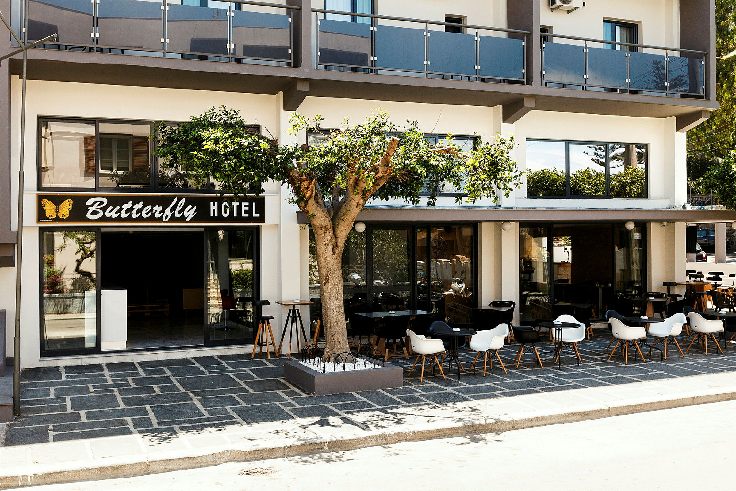 Forsiden av et hotell med skiltet "Butterfly Hotel", en uteplass med bord og stoler samt et tre foran bygningen. (AI-generert tekst)