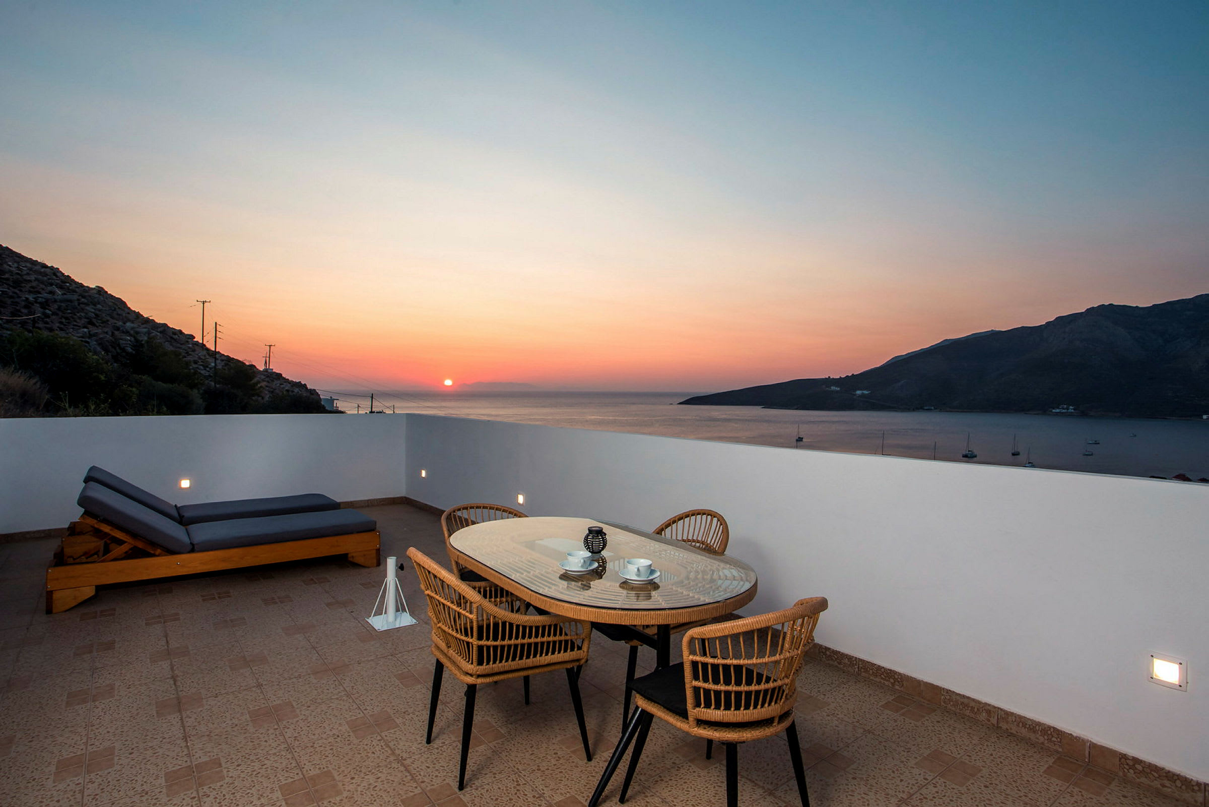 Utendørs terrasse med bord, stoler og solseng foran havutsikt ved solnedgang. (AI-generert tekst)