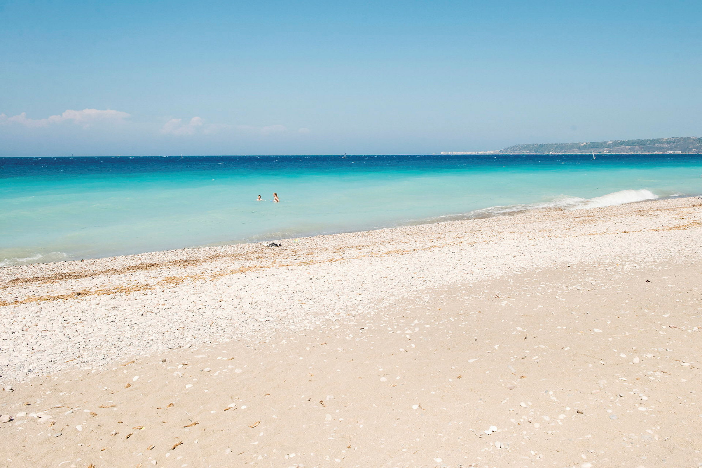 Hvit rullesteinsstrand ved havet med klart blått vann og to personer som bader. (AI-generert tekst)