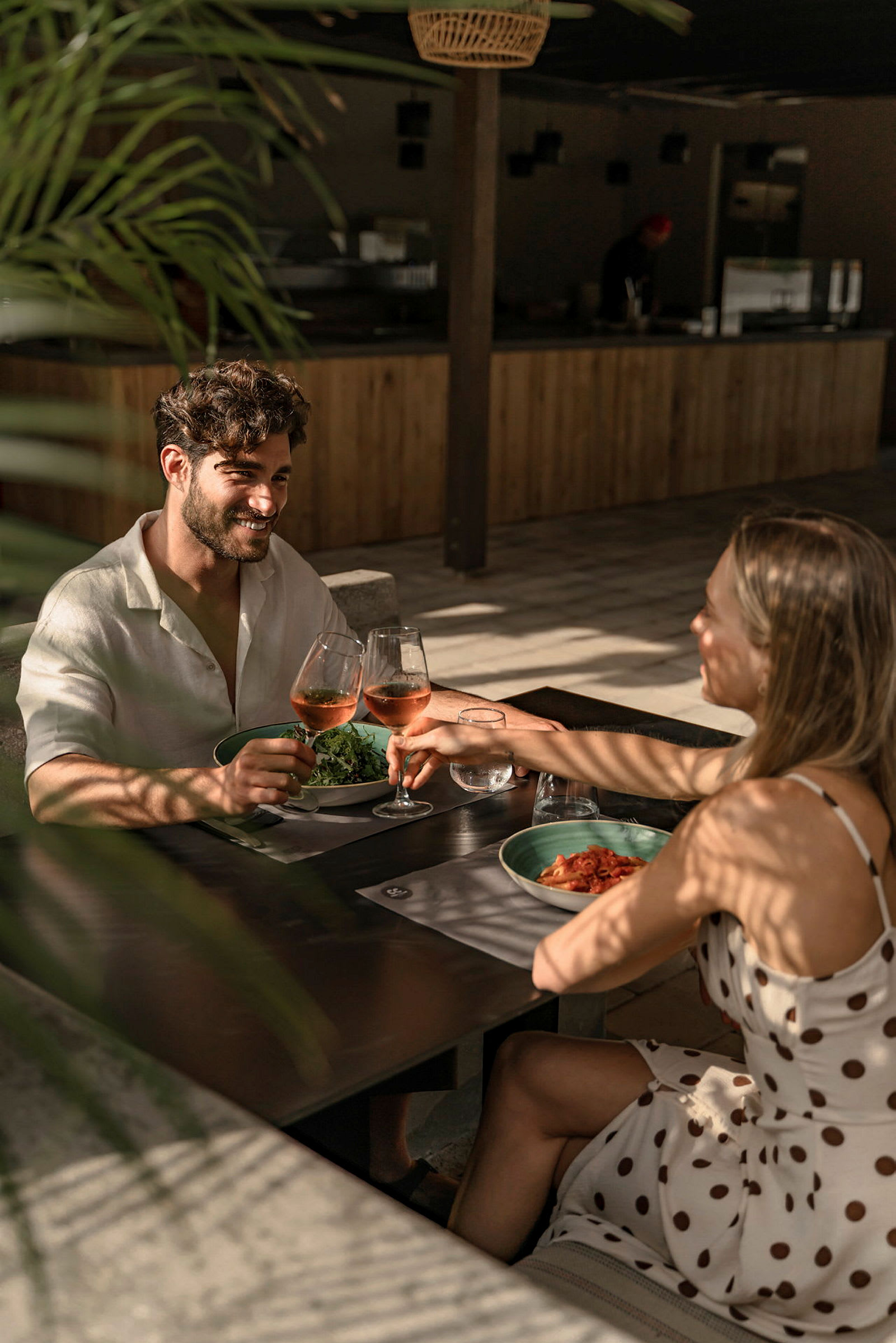 To personer skåler med glass som inneholder rosévin ved et bord på en solfylt restaurant. (AI-generert tekst)