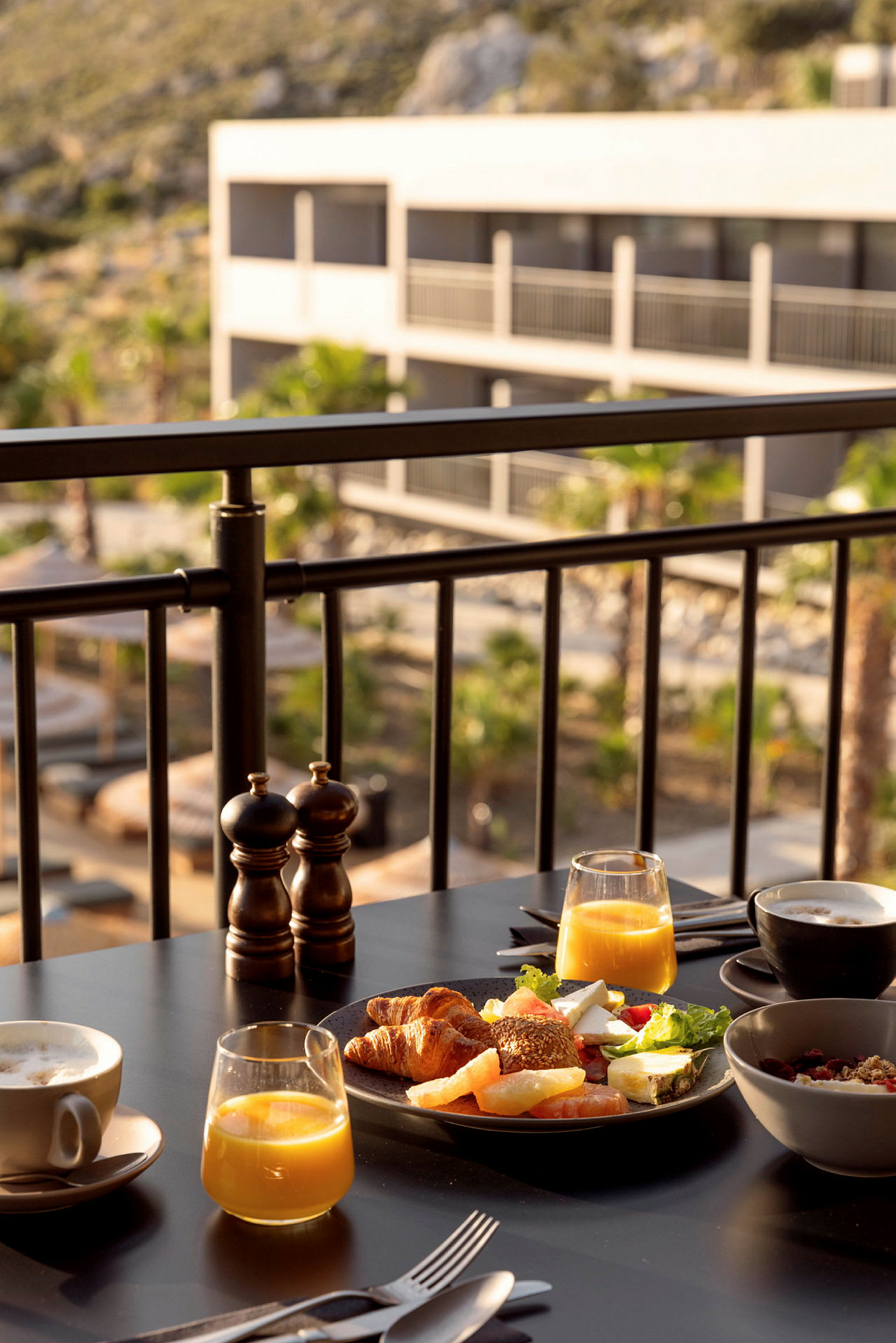 Frokost med juice, kaffe og forskjellige brød servert på et bord på et soldekk med utsikt over en hotellbygning. (AI-generert tekst)