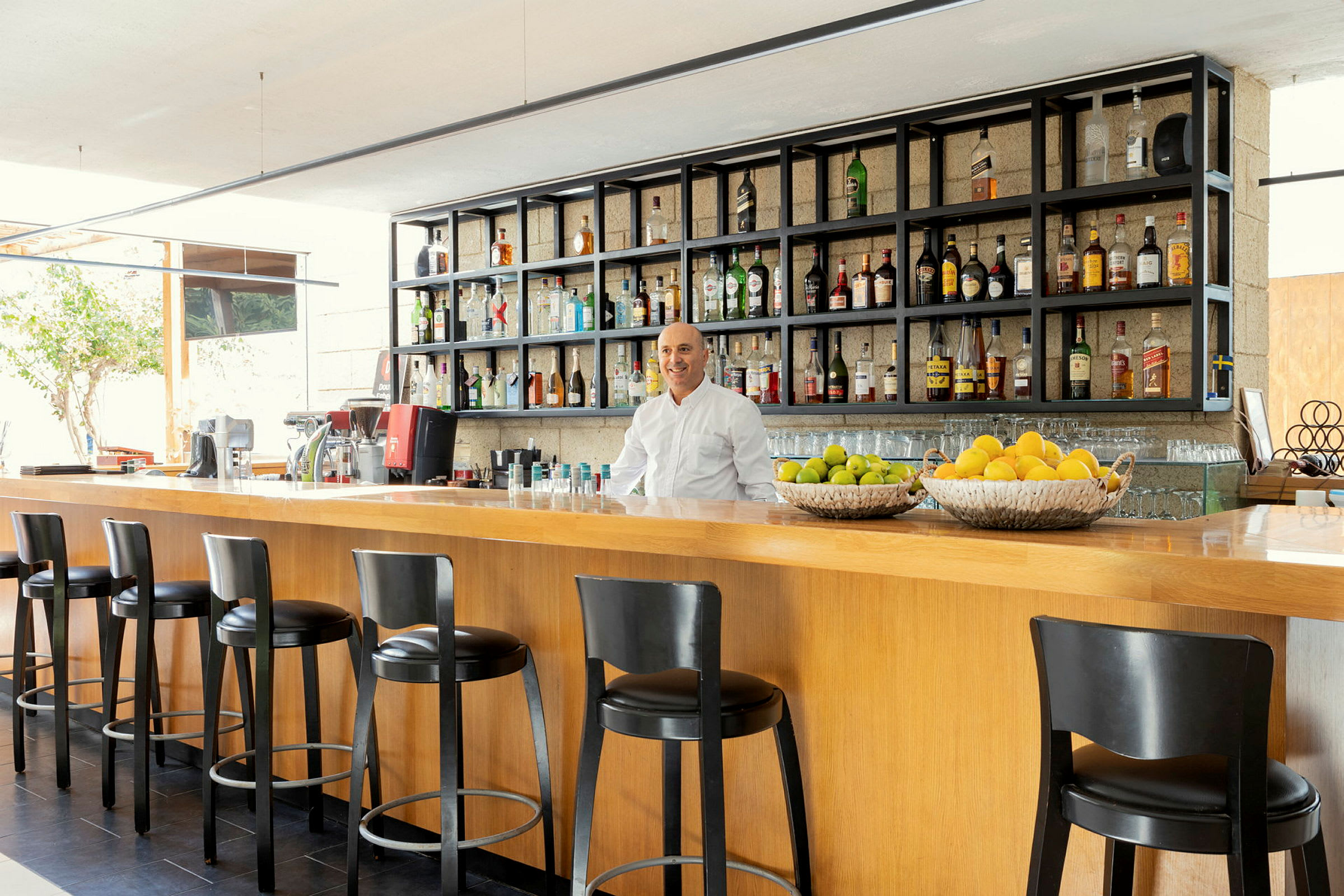 En moderne bar med høye stoler ved en trebardisk, hyller fylt med flasker og fruktkurver med sitrusfrukter. (AI-generert tekst)