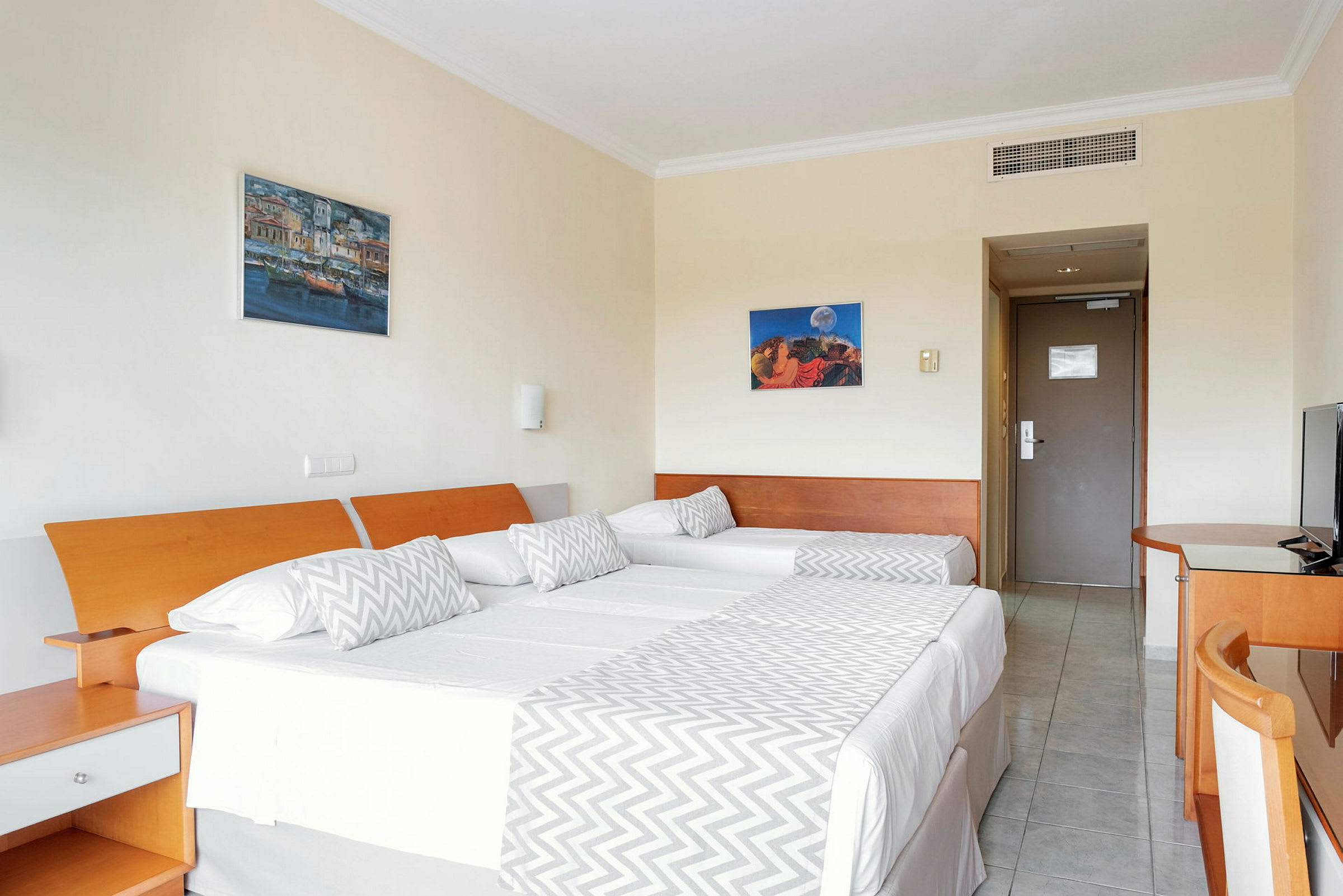 Bilde av et hotellrom med to enkeltsenger satt sammen, dekorert med hvite laken og grå sikksakk-mønstrede sengetepper. Bilder henger på veggen over sengene, og en dør samt møbler er synlige. (AI-generert tekst)