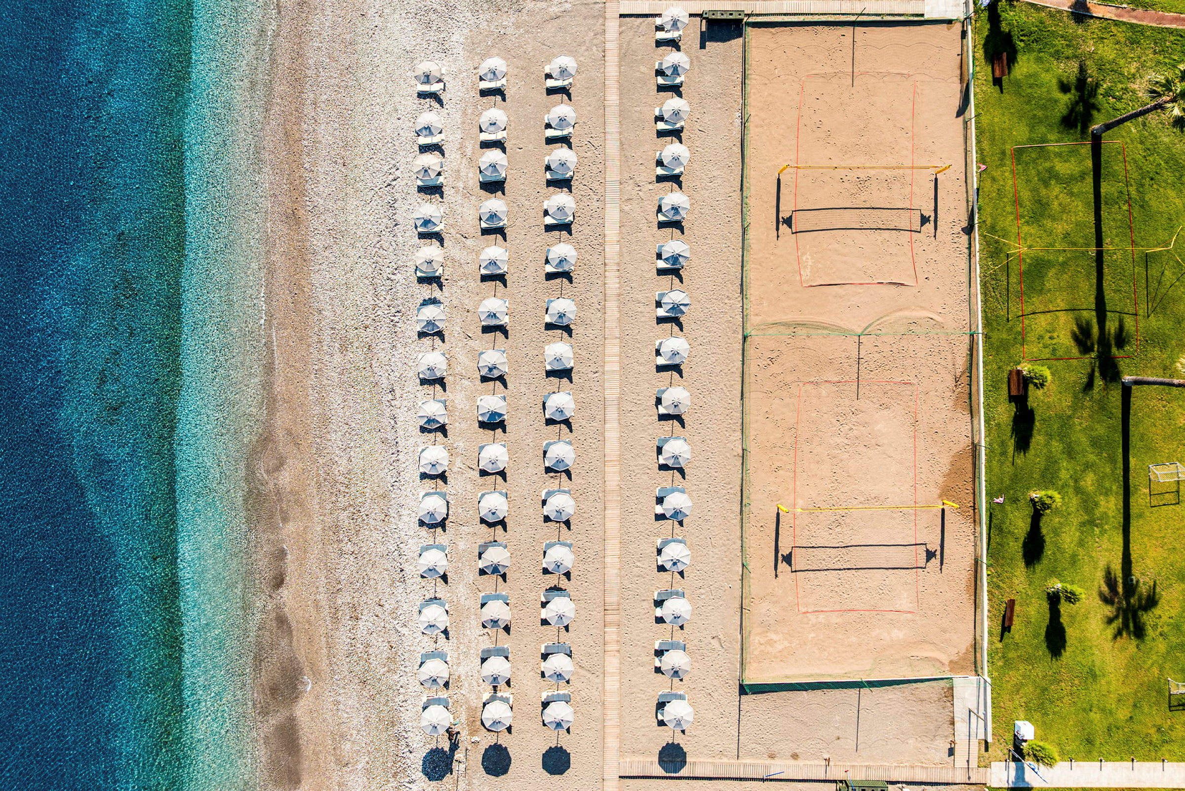 Luftfoto av en strand med parasoller oppstilt i rader, en volleyballbane på sand ved siden, og en gressplen med palmer og sitteplasser. (AI-generert tekst)