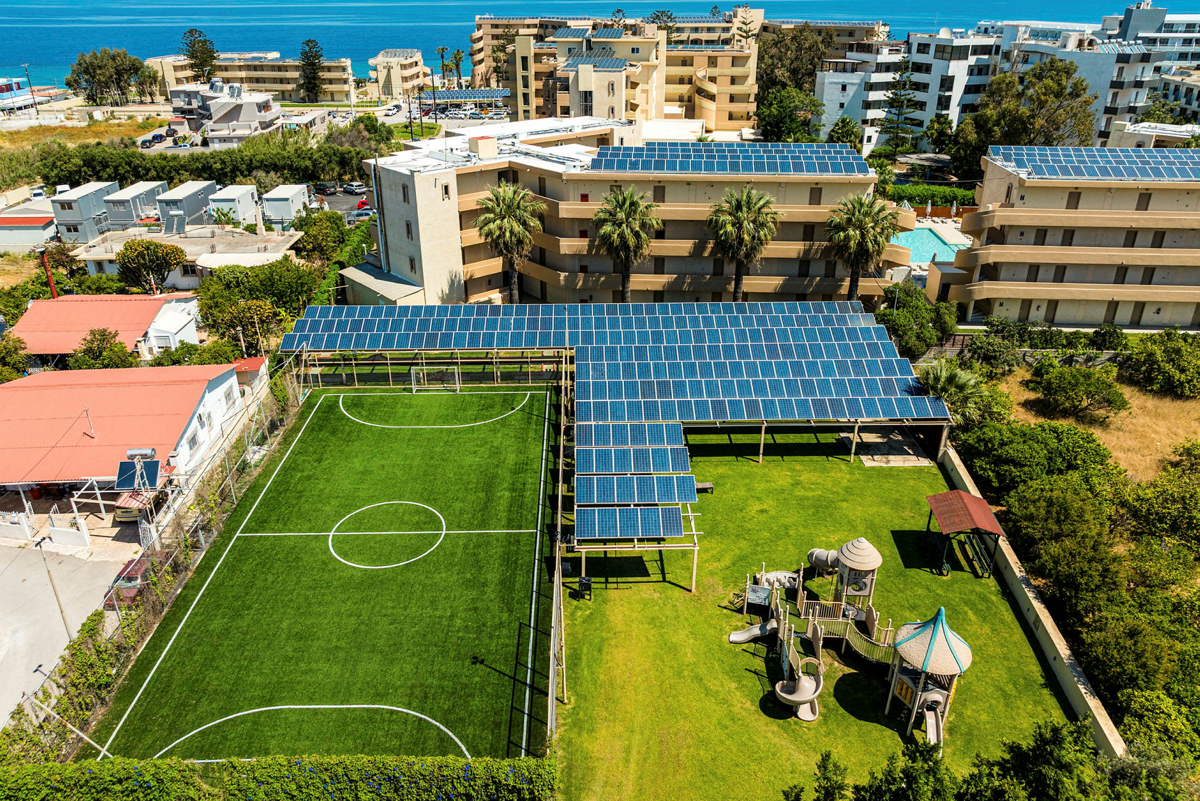 Bilde av et solfylt område med en grønn fotballbane og en lekeplass ved siden av. Bak disse er bygninger med solcellepaneler på takene. (AI-generert tekst)