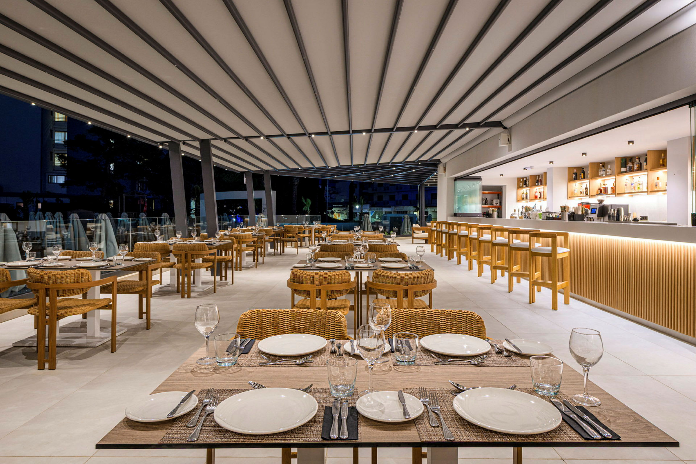 En moderne restaurant med dekkede bord og rottingstoler samt en lang bardisk i bakgrunnen. (AI-generert tekst)