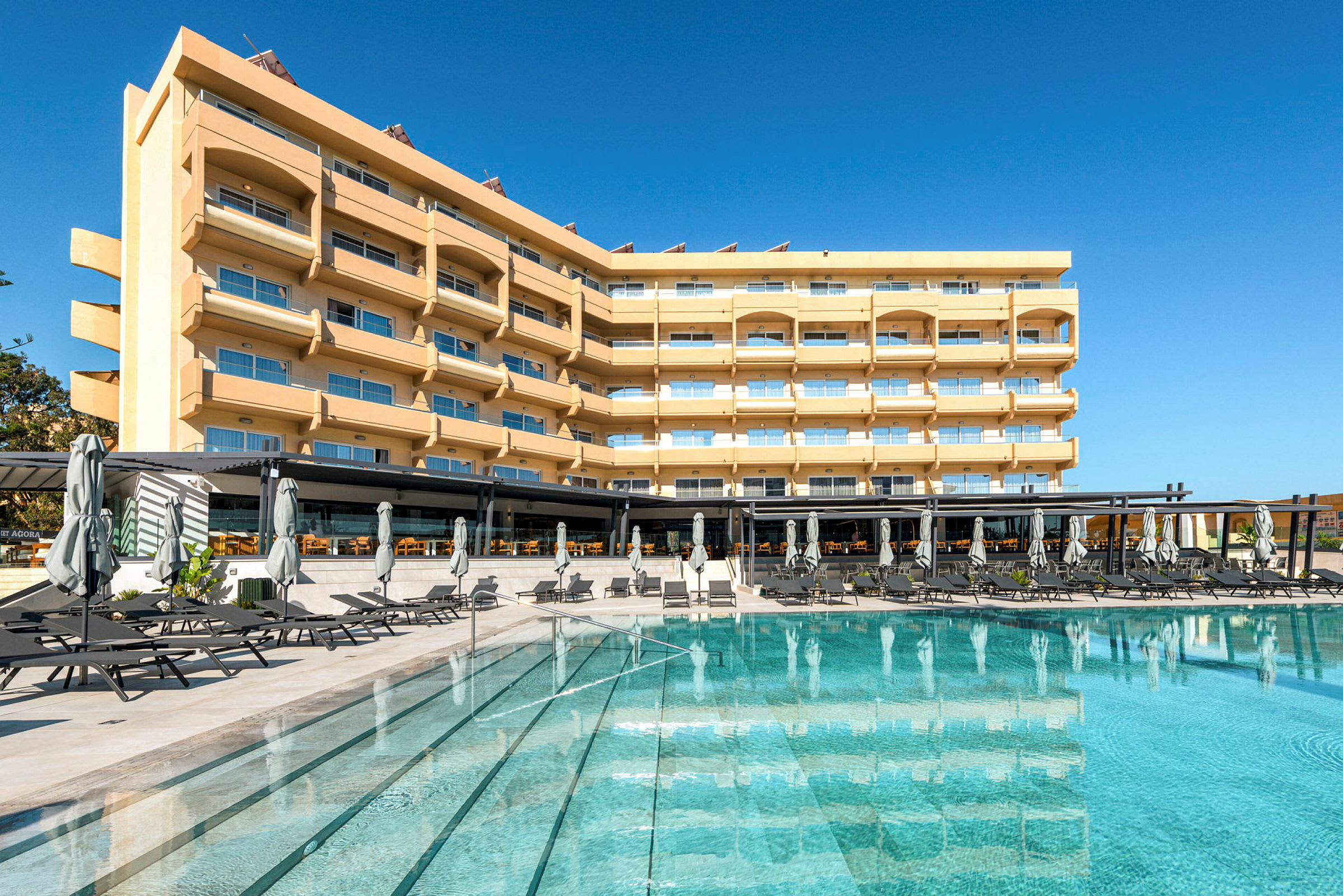 En hotellbygning med flere etasjer, balkonger og store vinduer, foran et svømmebasseng med solsenger og parasoller. (AI-generert tekst)