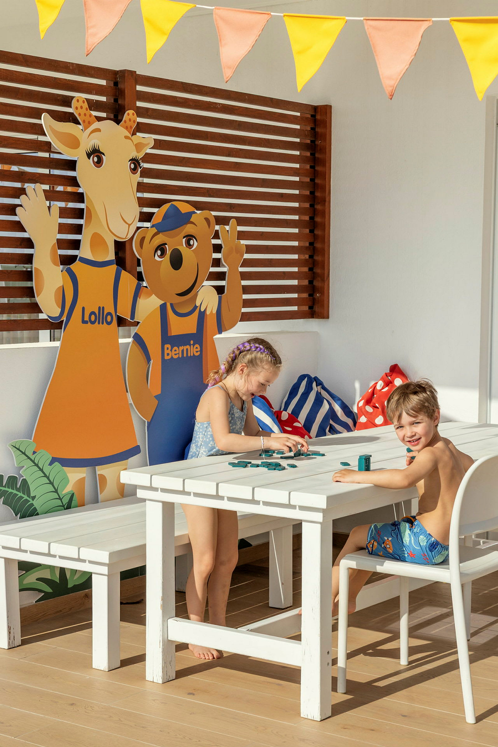 To barn sitter ved et utendørsbord og leker med spillebrikker. I bakgrunnen sees cutouts av figurene Lollo og Bernie. (AI-generert tekst)
