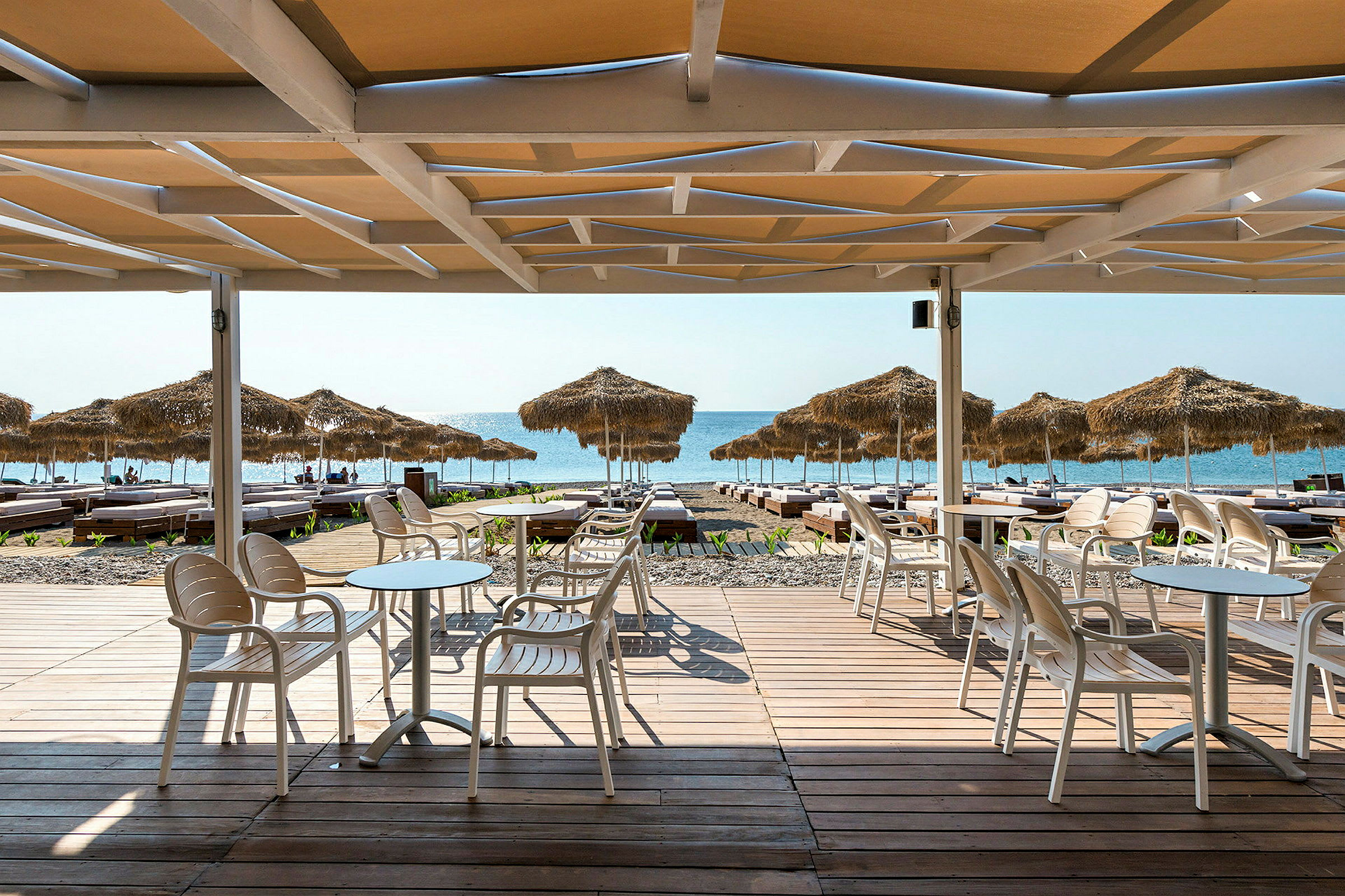 Strand med parasoller og solsenger, sett fra en treterrasse ved en uteservering. (AI-generert tekst)