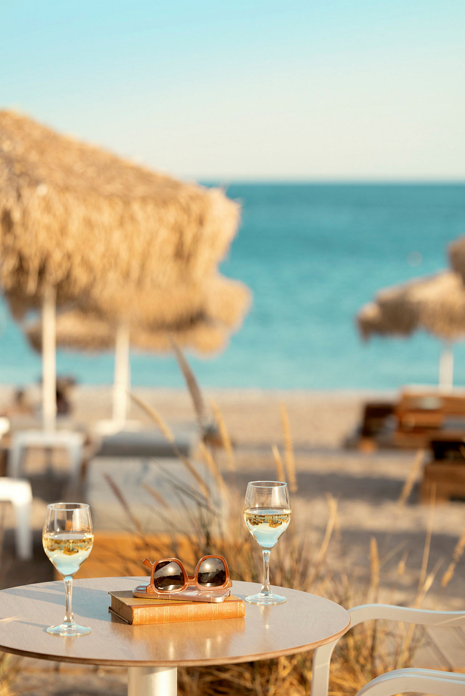 Bord med to glass hvitvin, en bok og solbriller ved en strand med parasoller og hav i bakgrunnen. (AI-generert tekst)