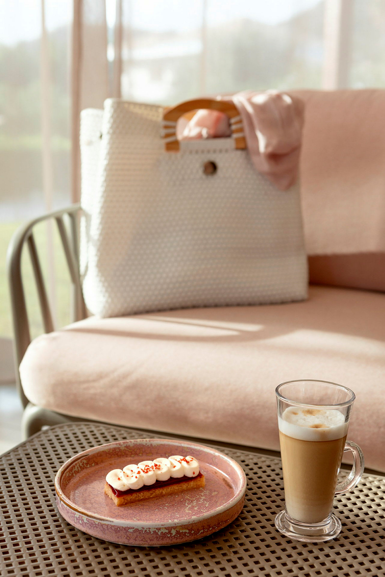 En beige veske med trehåndtak på en rosa sittemøbel med et glass latte og en dessert på en tallerken i forgrunnen. (AI-generert tekst)
