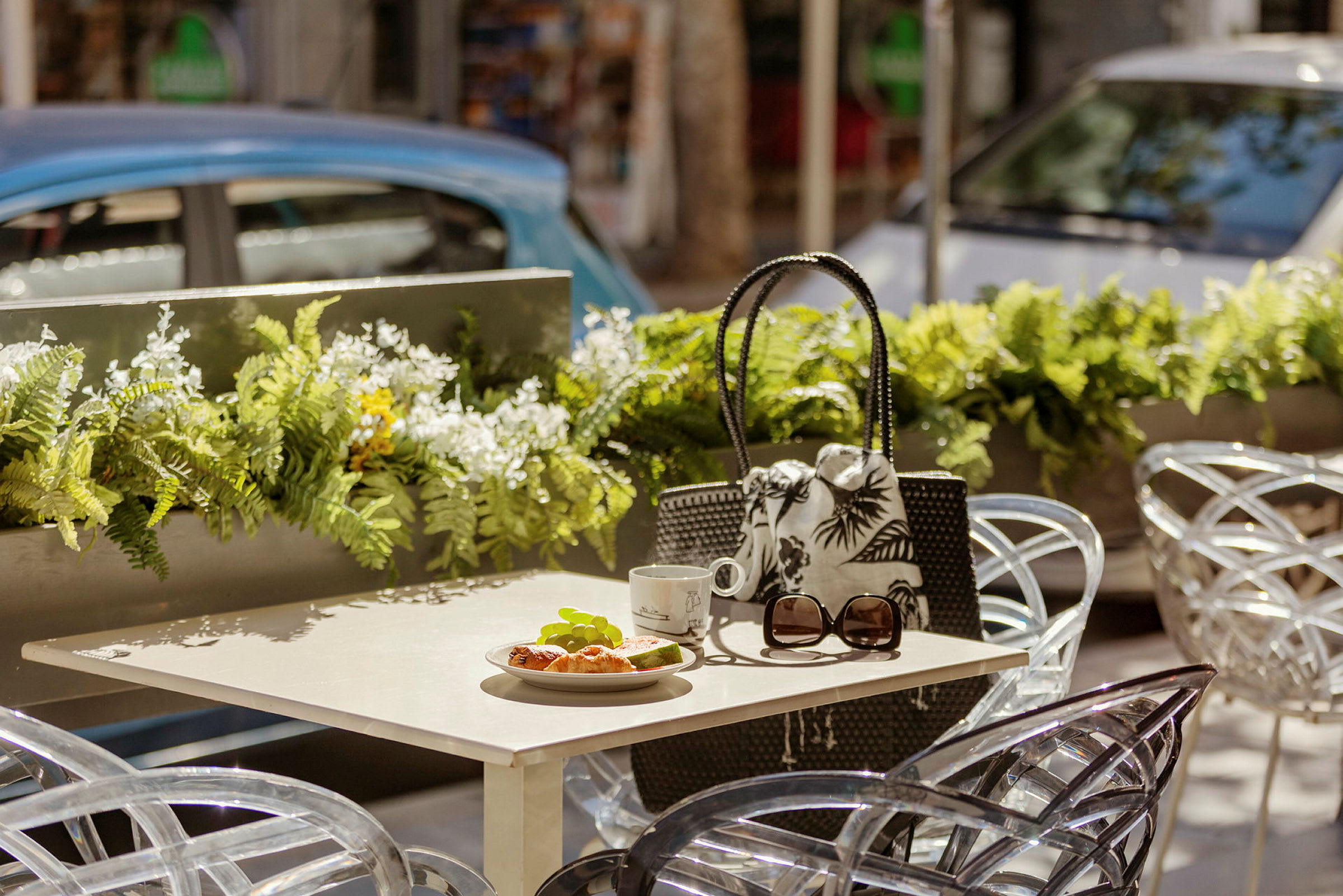 Bord med croissant, kopp og solbriller på uteservering med blomsterdekorasjoner. (AI-generert tekst)