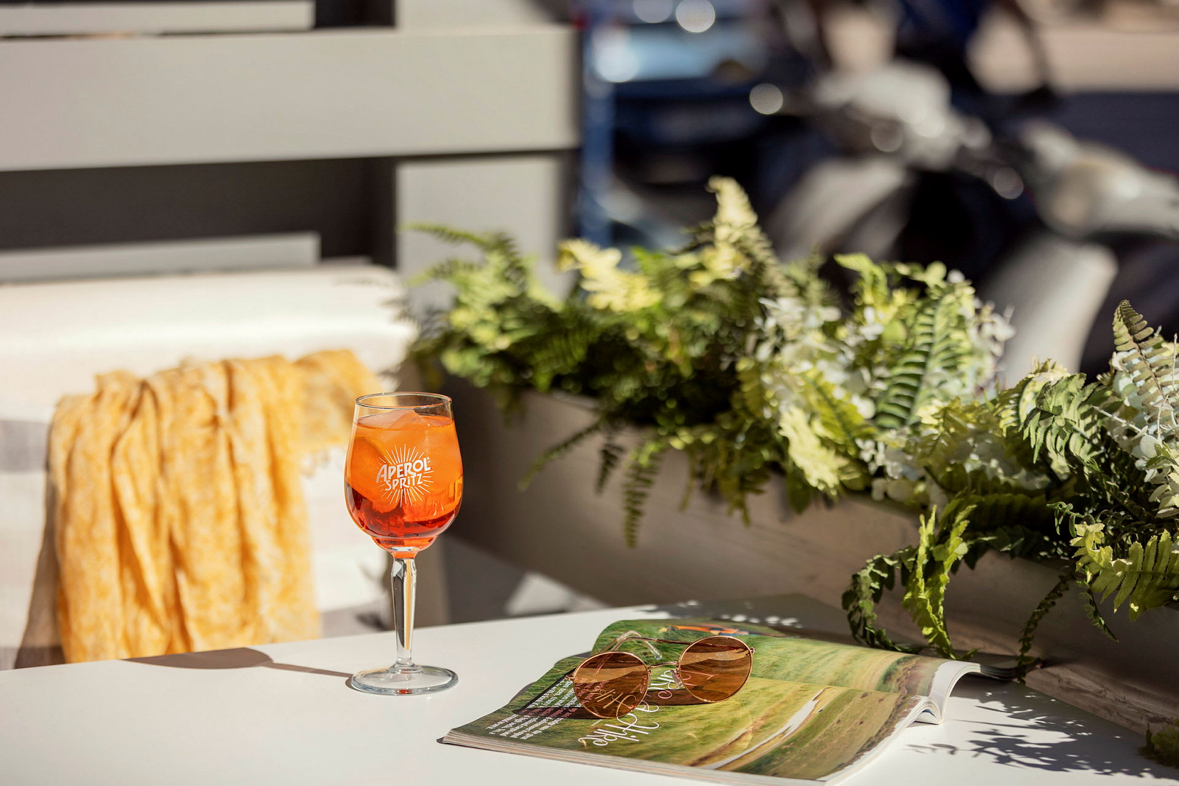 Et glass Aperol Spritz står på et bord ved siden av en solbrille og et åpent magasin. I bakgrunnen ses planter og deler av en balkong. (AI-generert tekst)