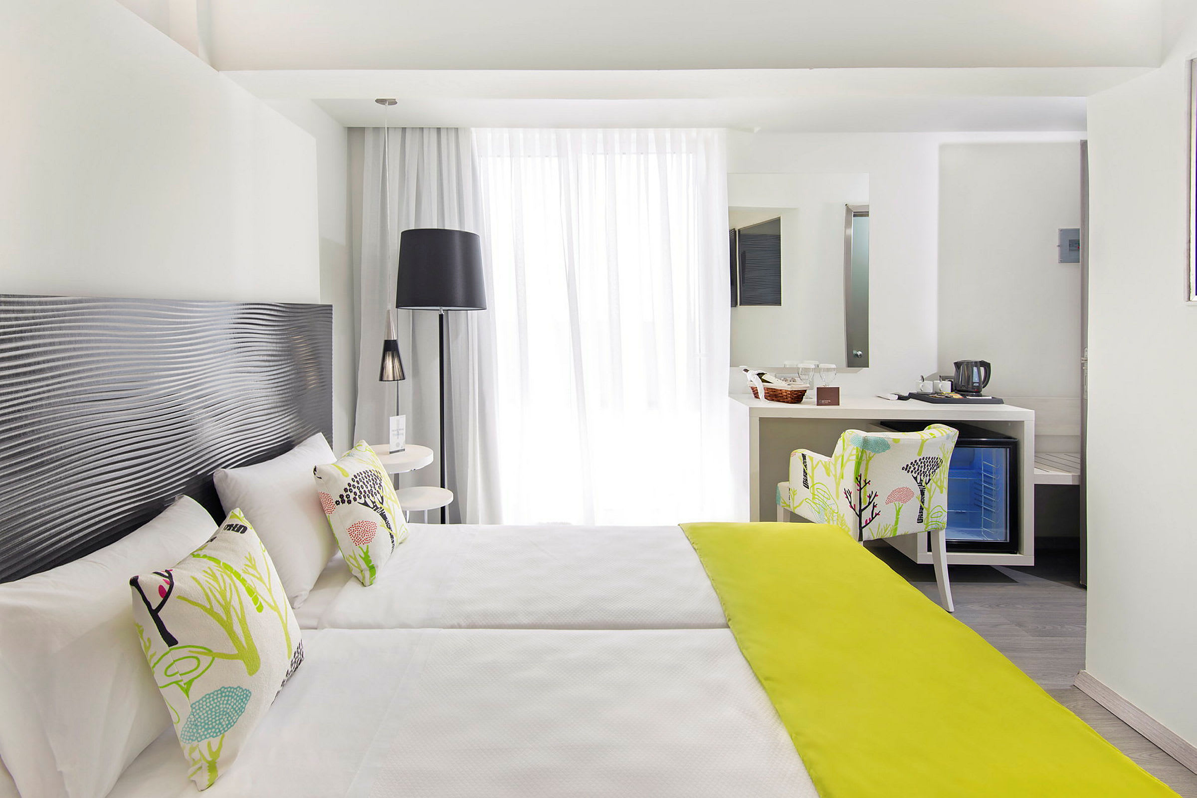 Et lyst hotellrom med to enkeltsenger, dekorative puter og et limegrønt sengeteppe, samt en sitteplass og et skrivebord med speil. (AI-generert tekst)