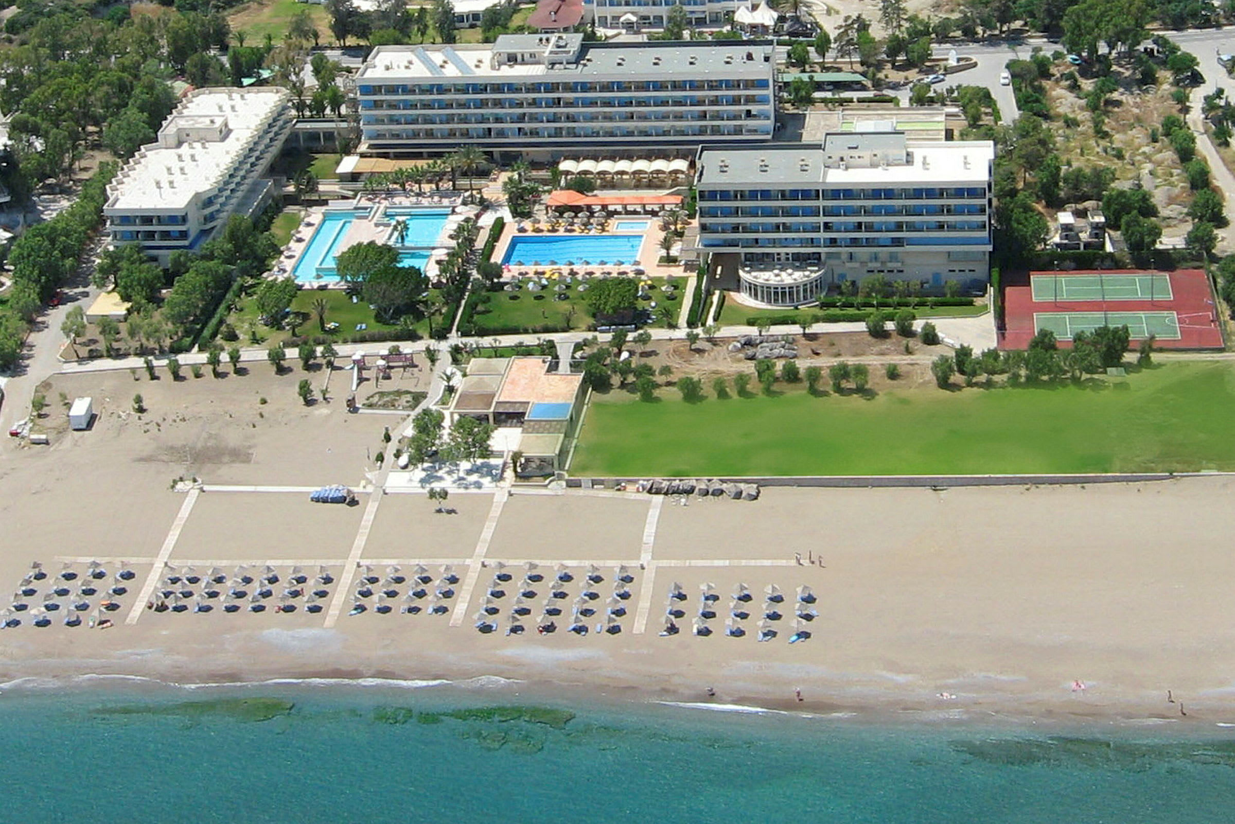 Luftperspektiv av et hotellkompleks med bassenger, grøntområder og tennisbaner nær en sandstrand. (AI-generert tekst)