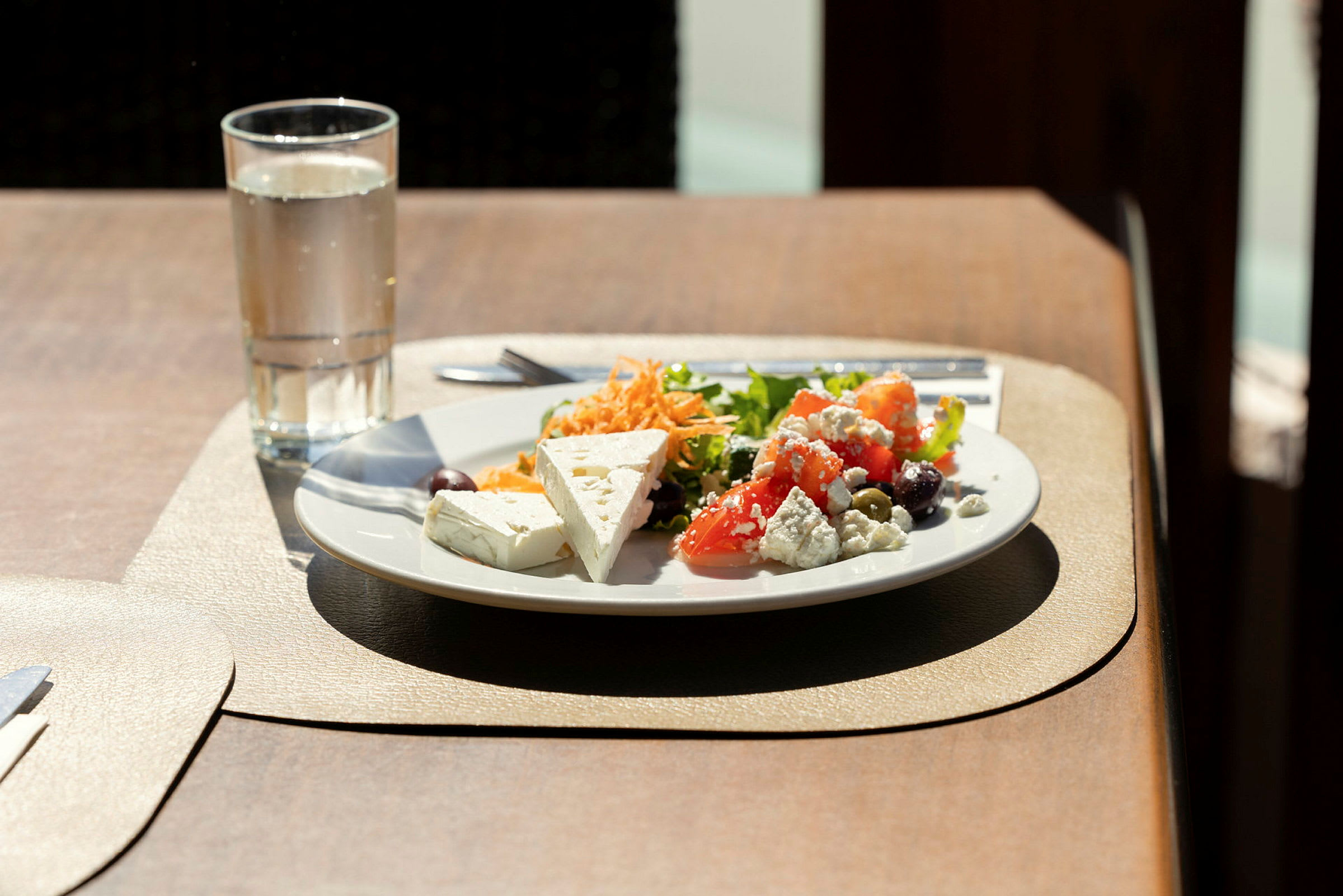 En tallerken med salat, grønnsaker og fetaost samt et glass vann på et bord. (AI-generert tekst)