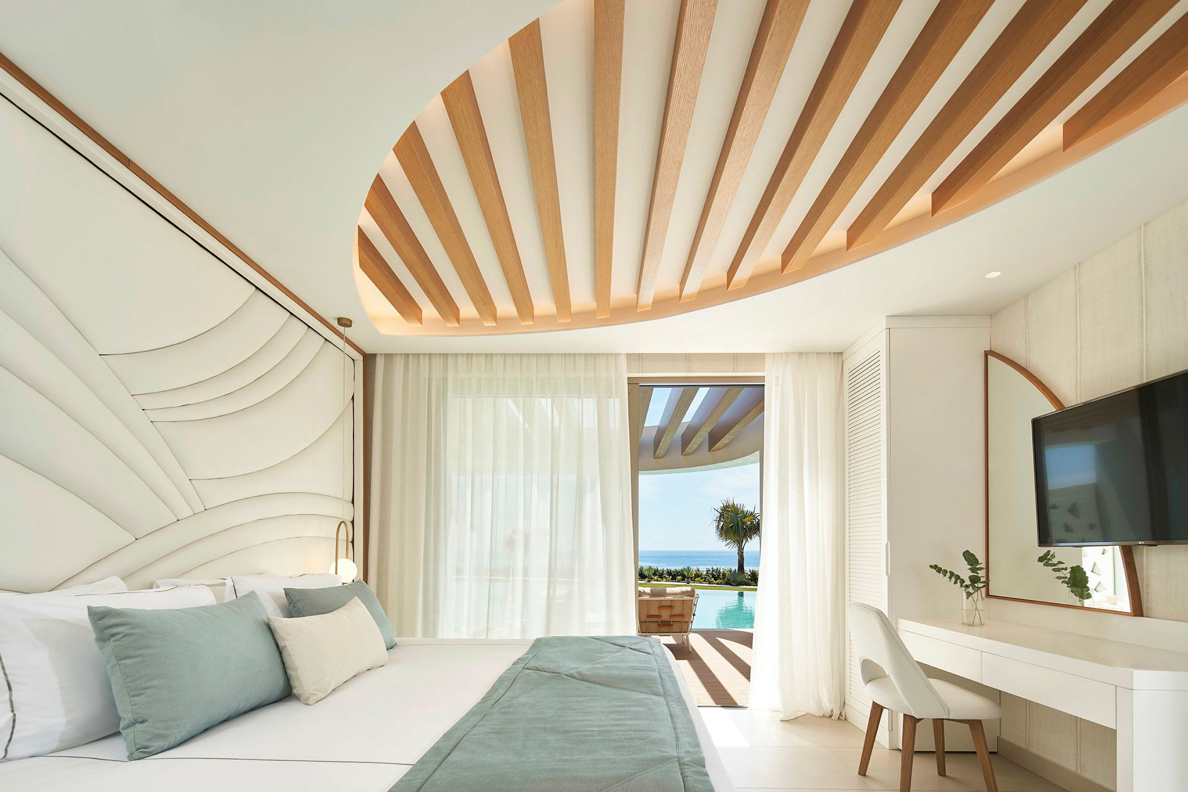 Et moderne hotellrom med en stor seng, designede takdetaljer og utsikt over et basseng og havet gjennom en glassdør. (AI-generert tekst)