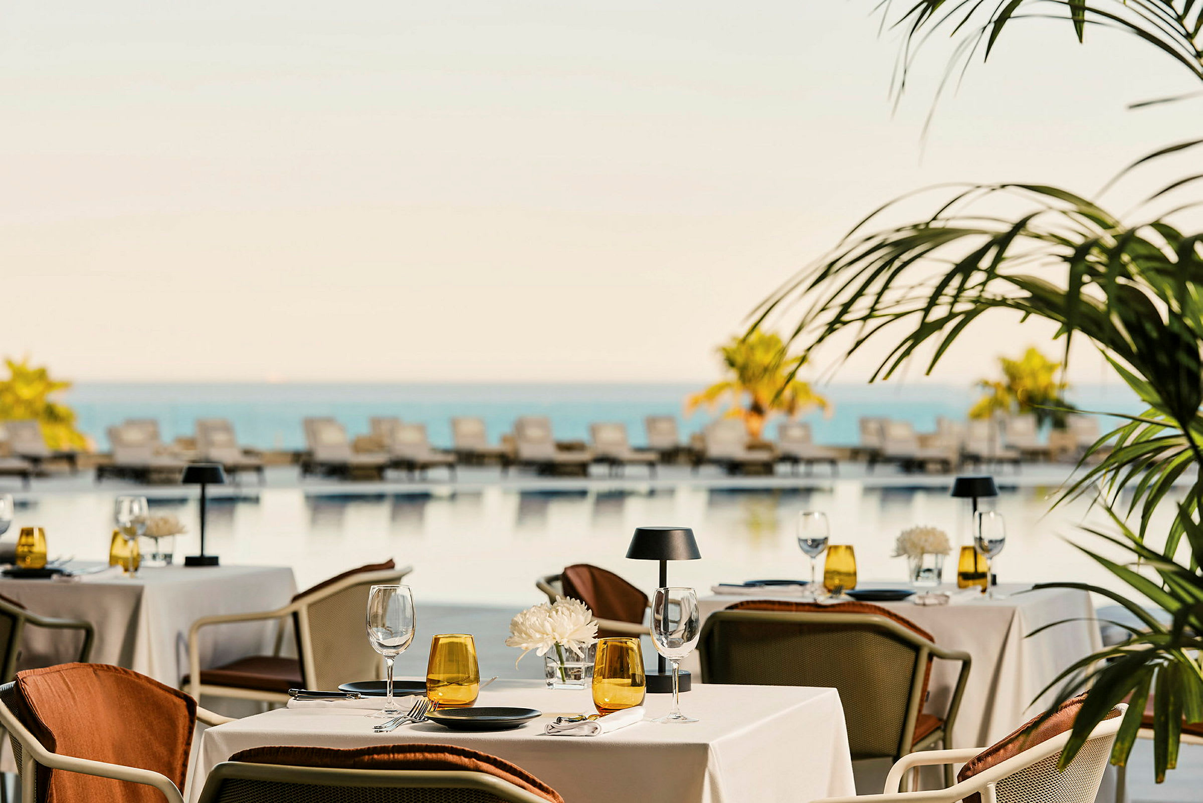 En restaurant med dekkede bord ved et basseng med solsenger og utsikt over havet. (AI-generert tekst)
