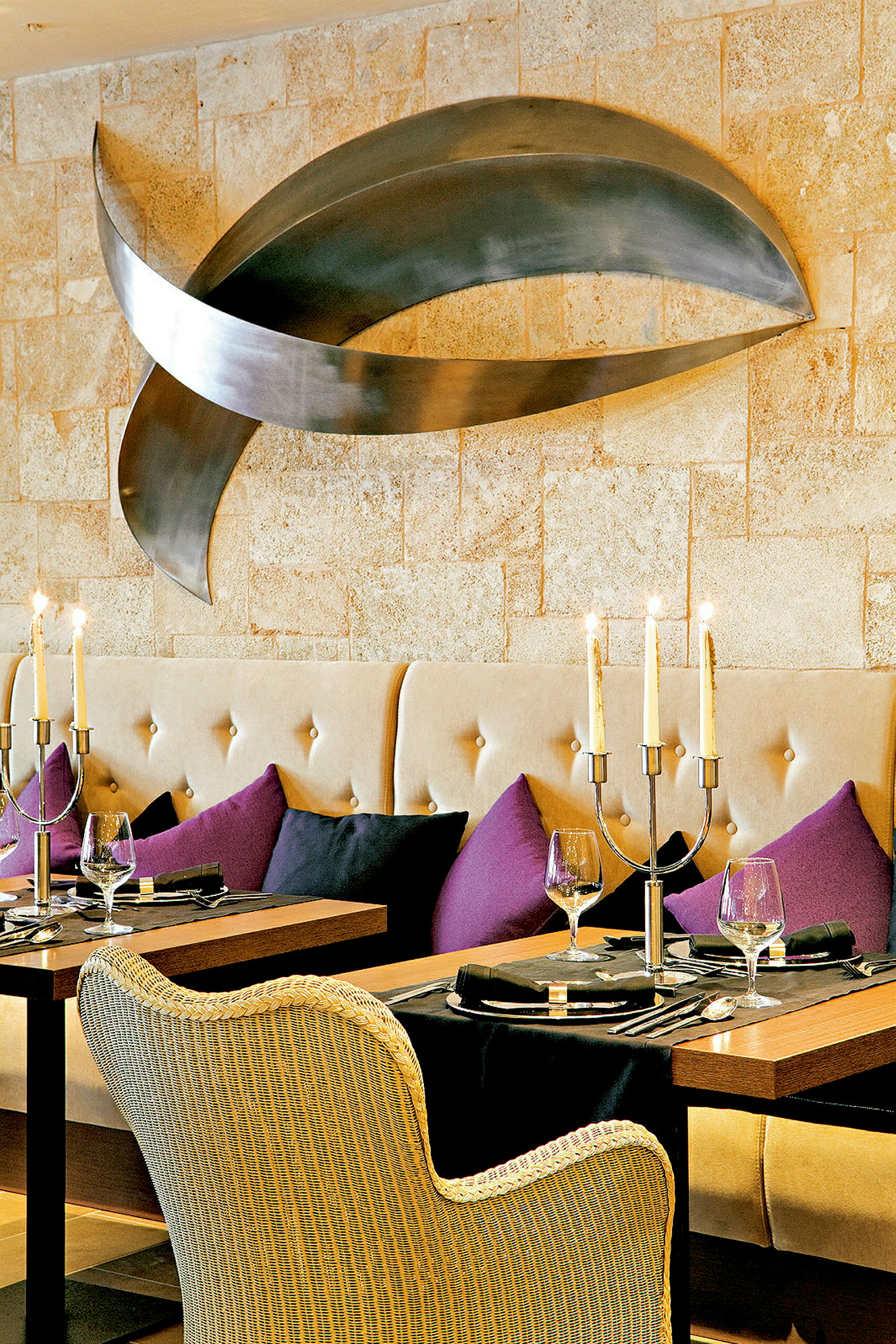 Et elegant restaurantbord med tallerkener, glass og lysestaker, omgitt av stoppede sofaer og fargede puter, og en moderne kunstinstallasjon på veggen. (AI-generert tekst)