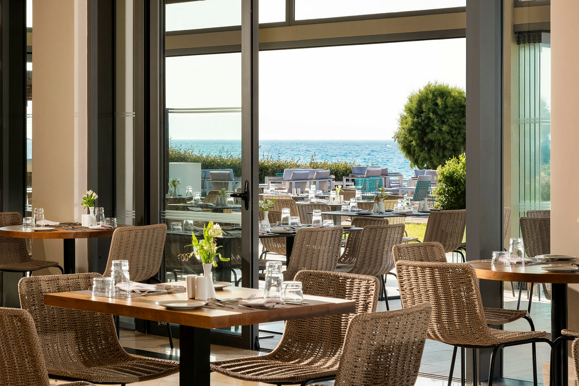 Utsikt over en moderne restaurant med flettestoler og bord, med en hage og havet i bakgrunnen. (AI-generert tekst)