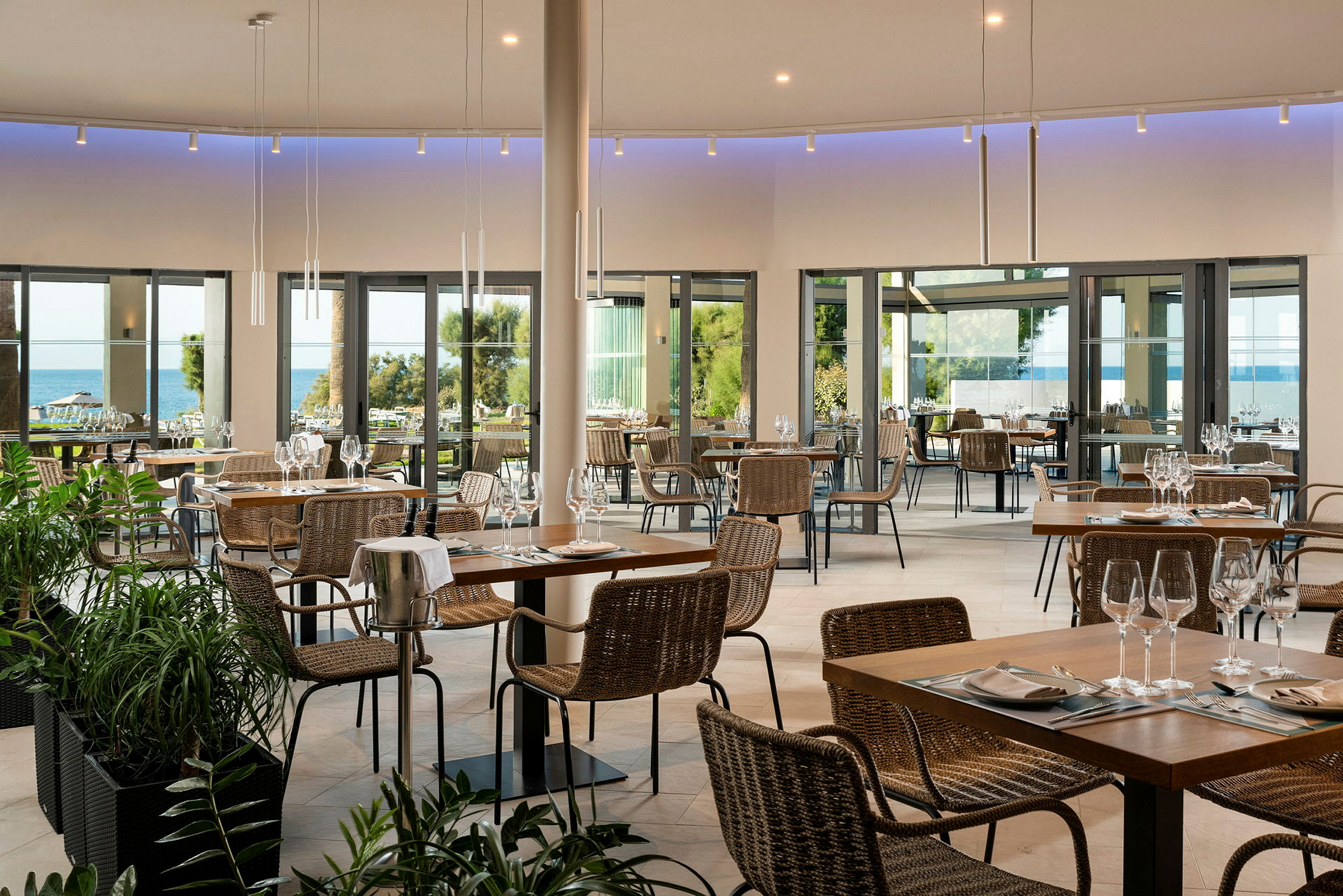 En restaurant med bord og stoler, store glassvinduer med utsikt over havet og grøntområder. (AI-generert tekst)