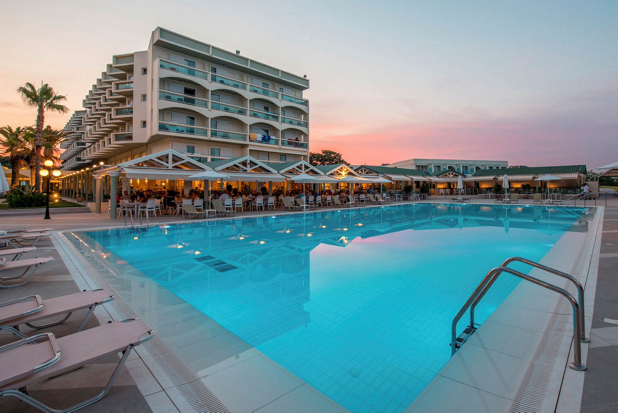 Hotellbygning med balkonger ved et stort svømmebasseng omgitt av solsenger og en terrasse med sitteplasser. (AI-generert tekst)