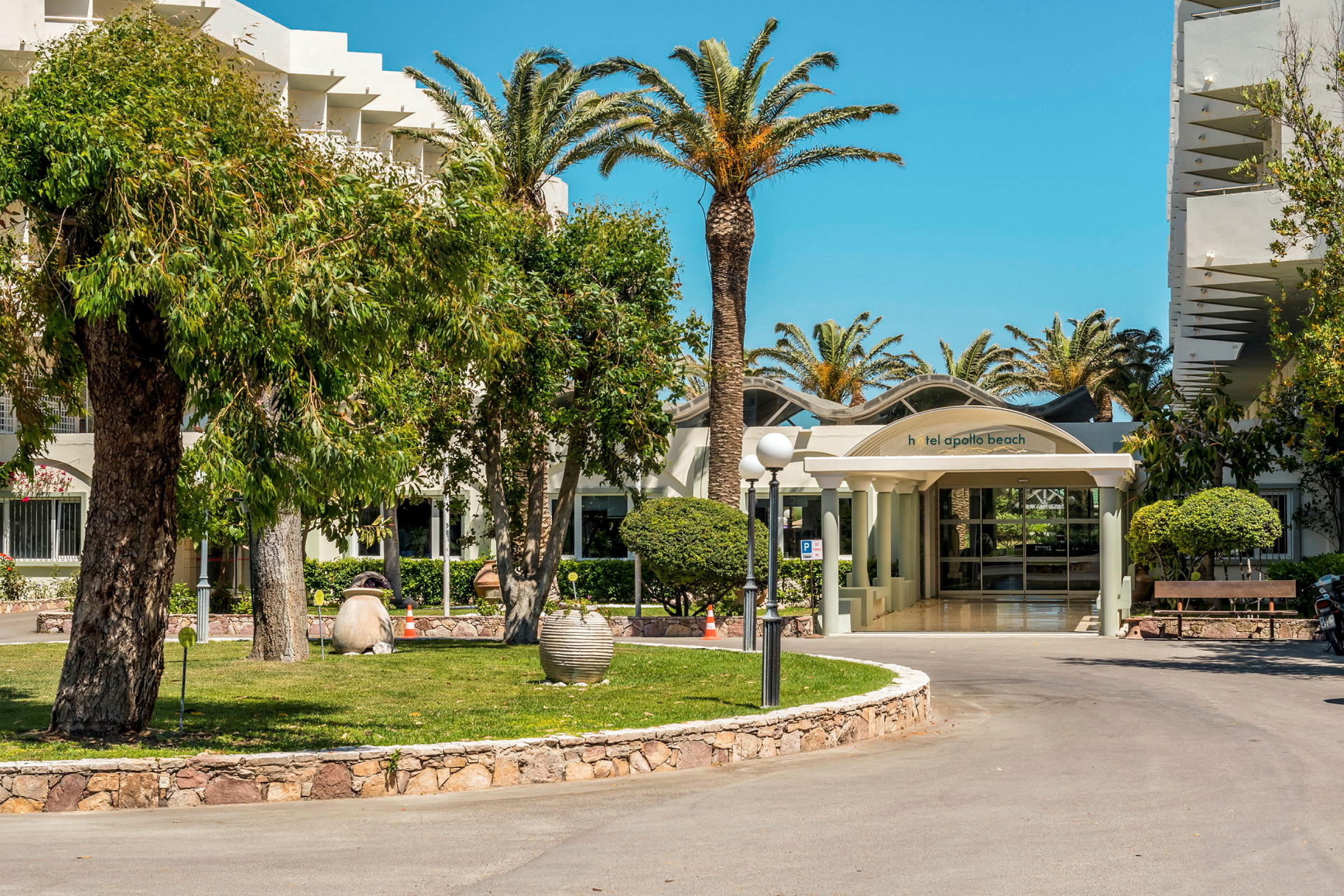 Inngangen til Hotel Apollo Beach omgitt av palmer og grøntområder. (AI-generert tekst)
