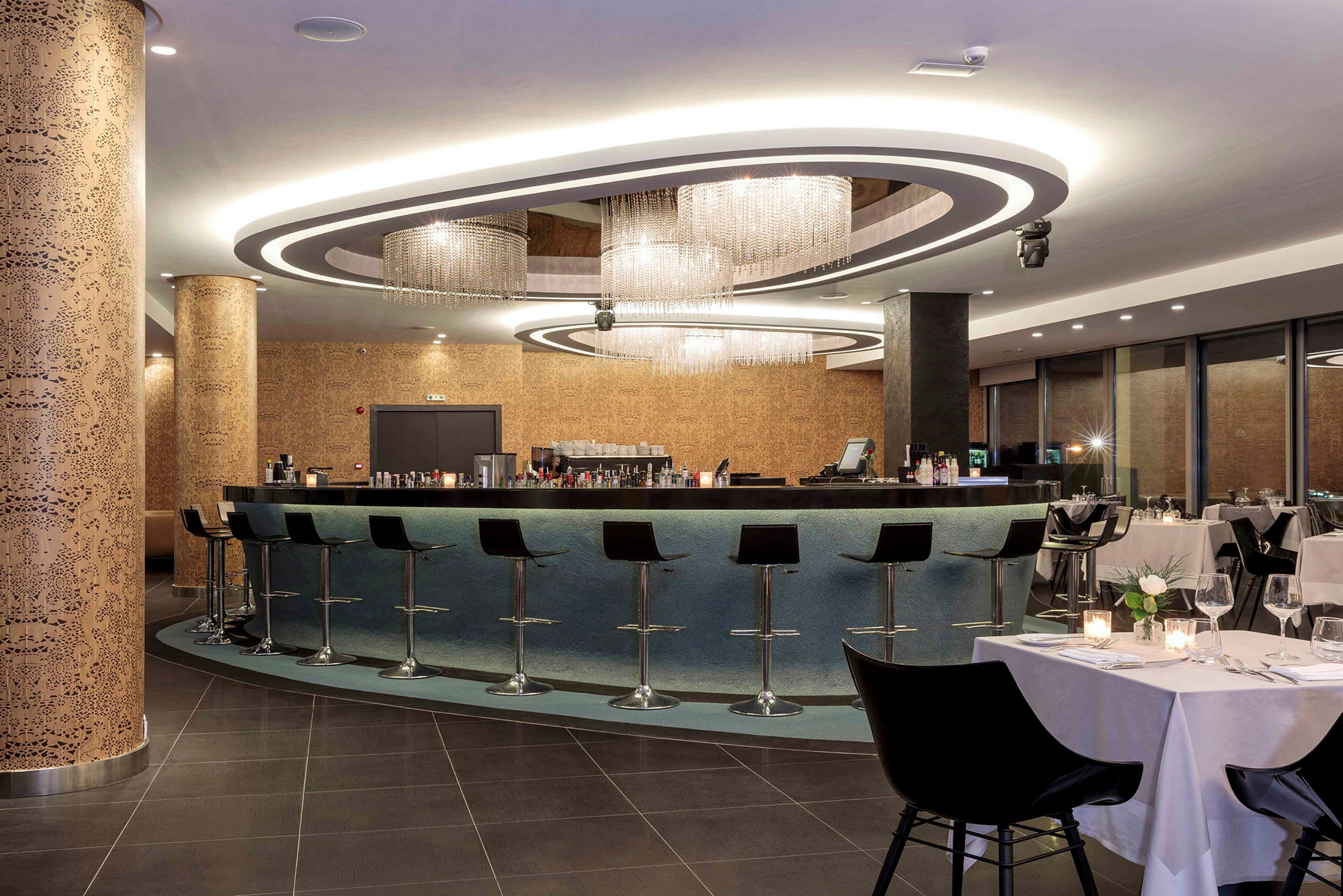 En moderne bar med høye stoler og elegant lysdesign, omgitt av en restaurant med dekkede bord. (AI-generert tekst)