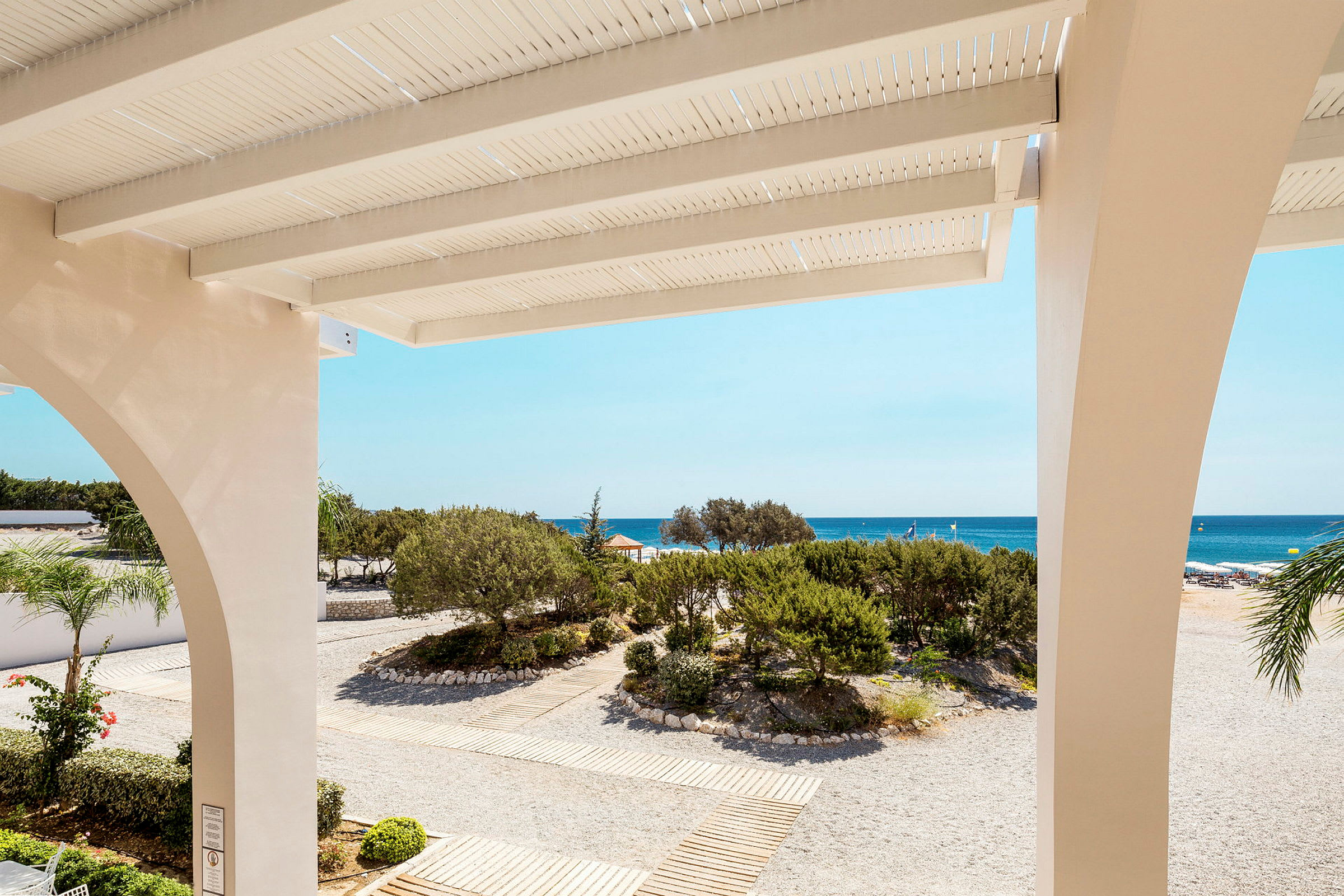 Utsikt fra en terrasse mot en strand med sand og grøntområder samt havet i bakgrunnen. (AI-generert tekst)