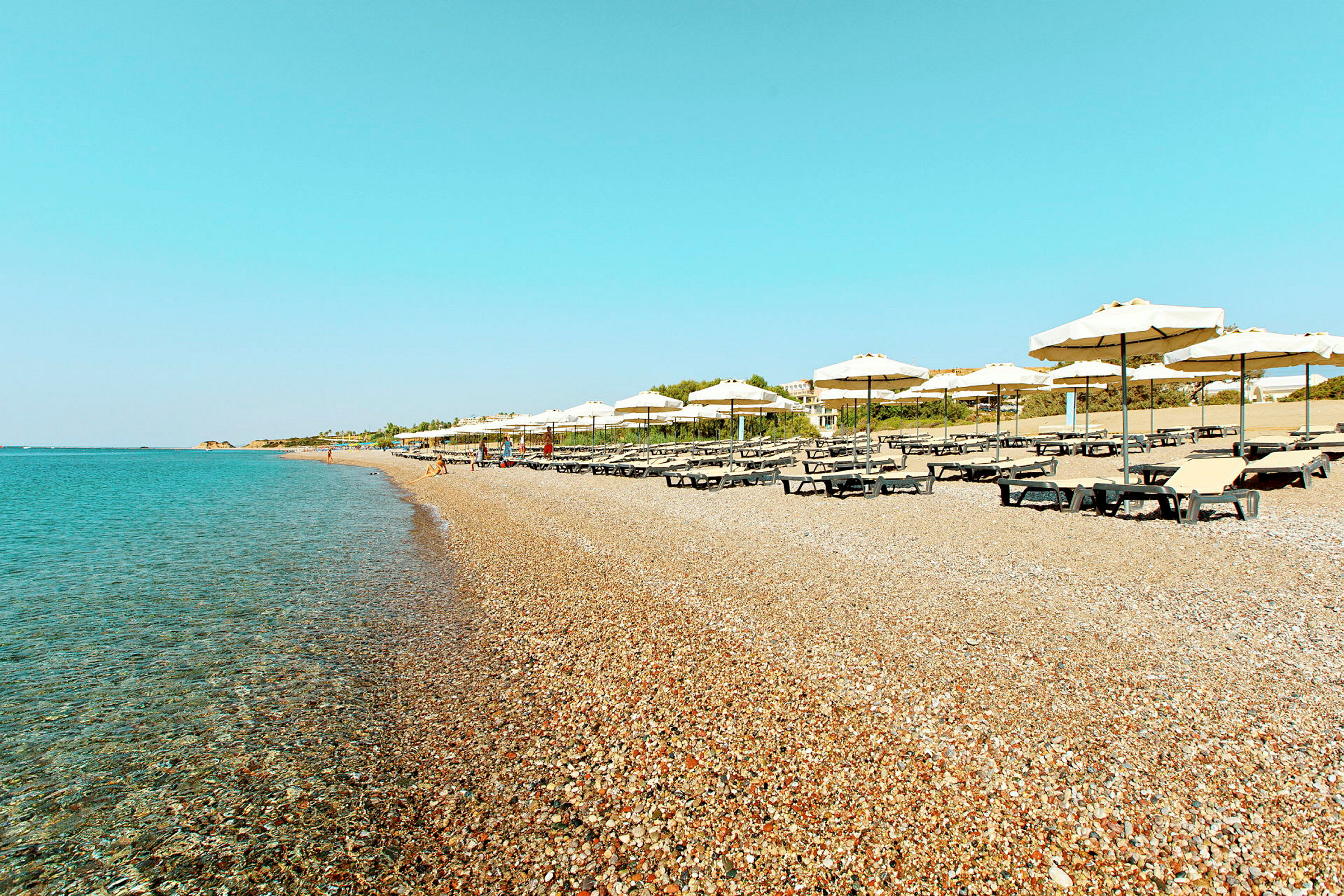 Et strandbilde med parasoller og solsenger på en steinete strand ved klart blått vann. (AI-generert tekst)