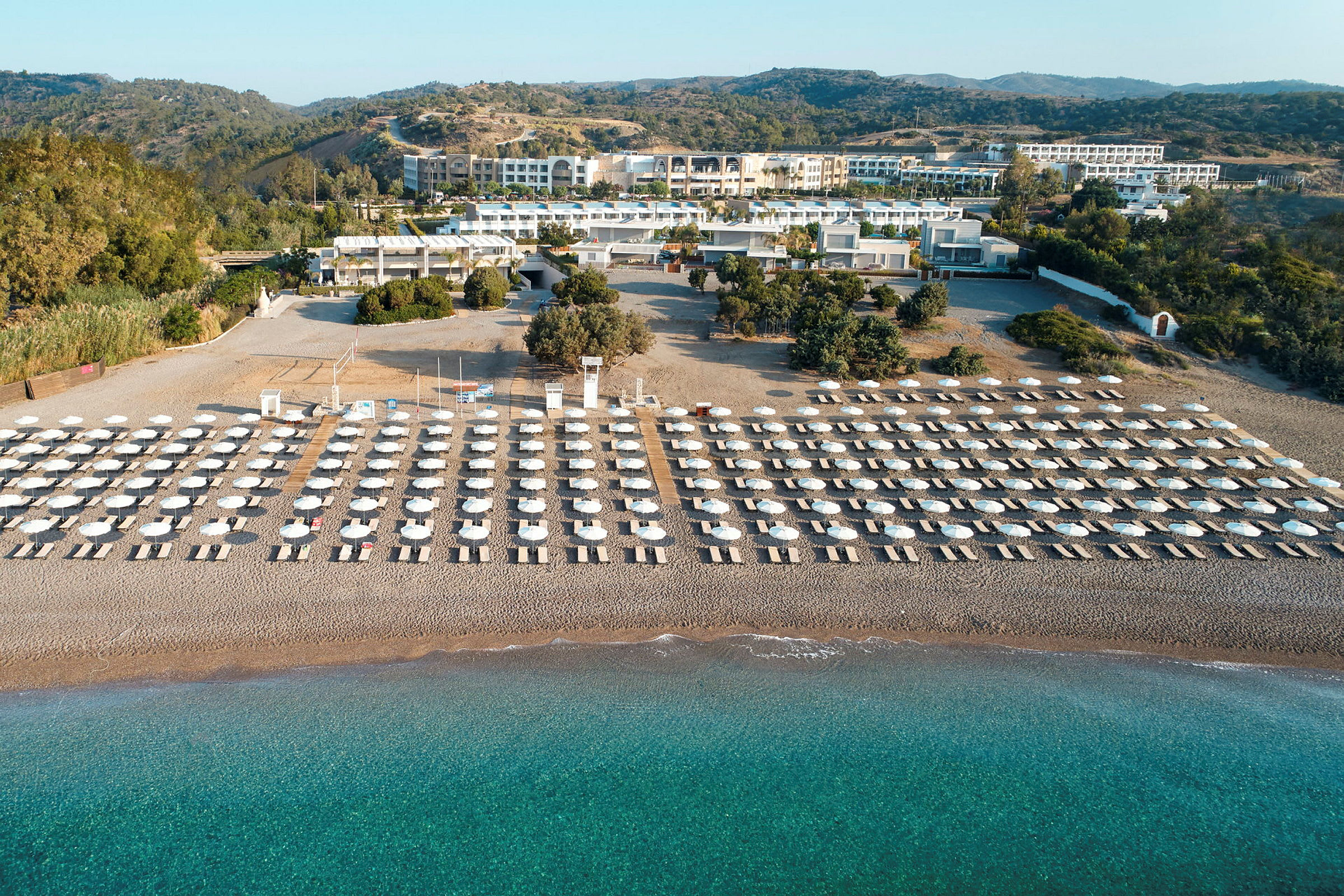 Solstoler og parasoller i flere rader på en sandstrand med turkis vann, omgitt av grøntområder og bygninger i bakgrunnen. (AI-generert tekst)