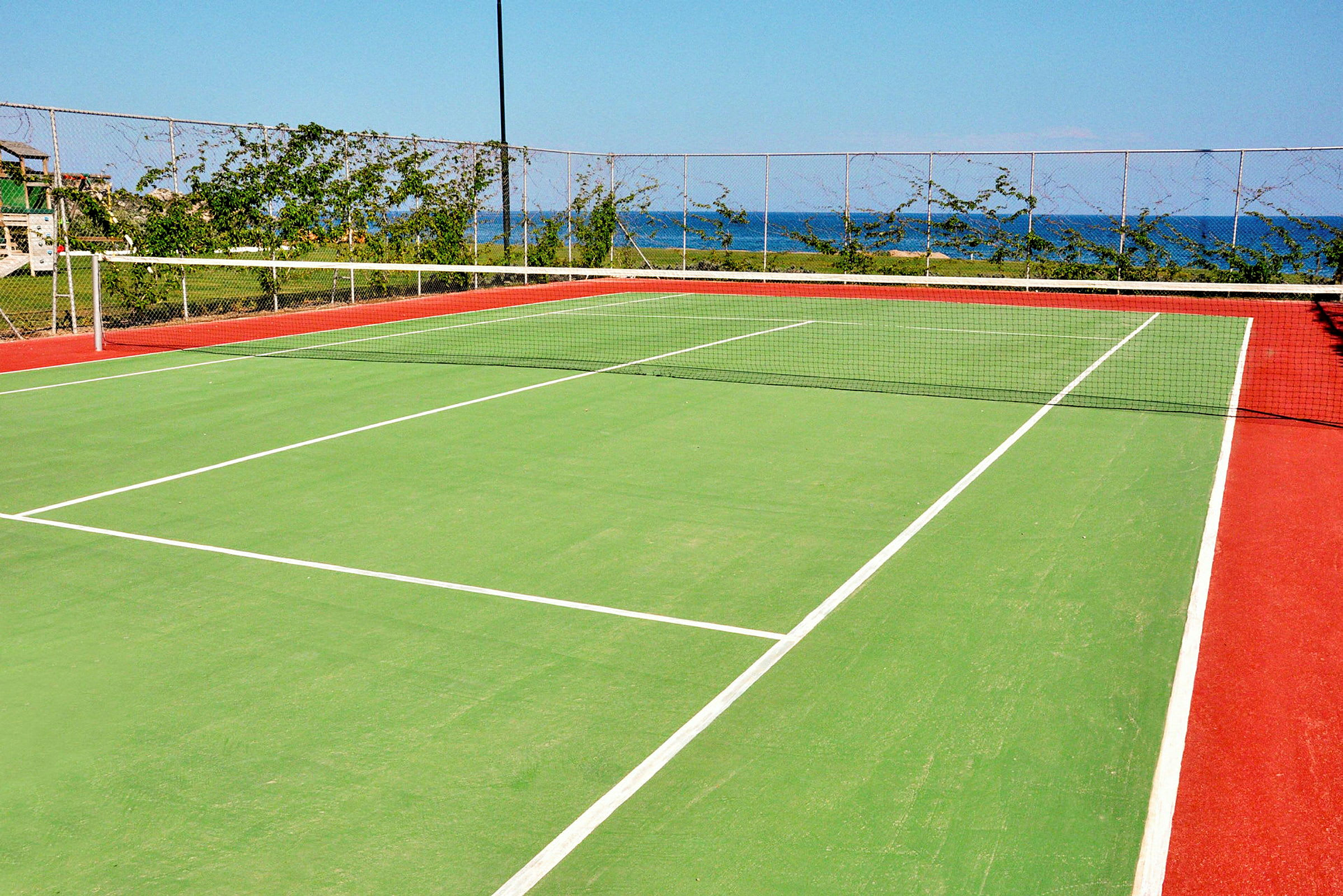 En tennisbane med grønn overflate og røde kanter, omgitt av et nettinggjerde og vegetasjon, med havet synlig i bakgrunnen. (AI-generert tekst)