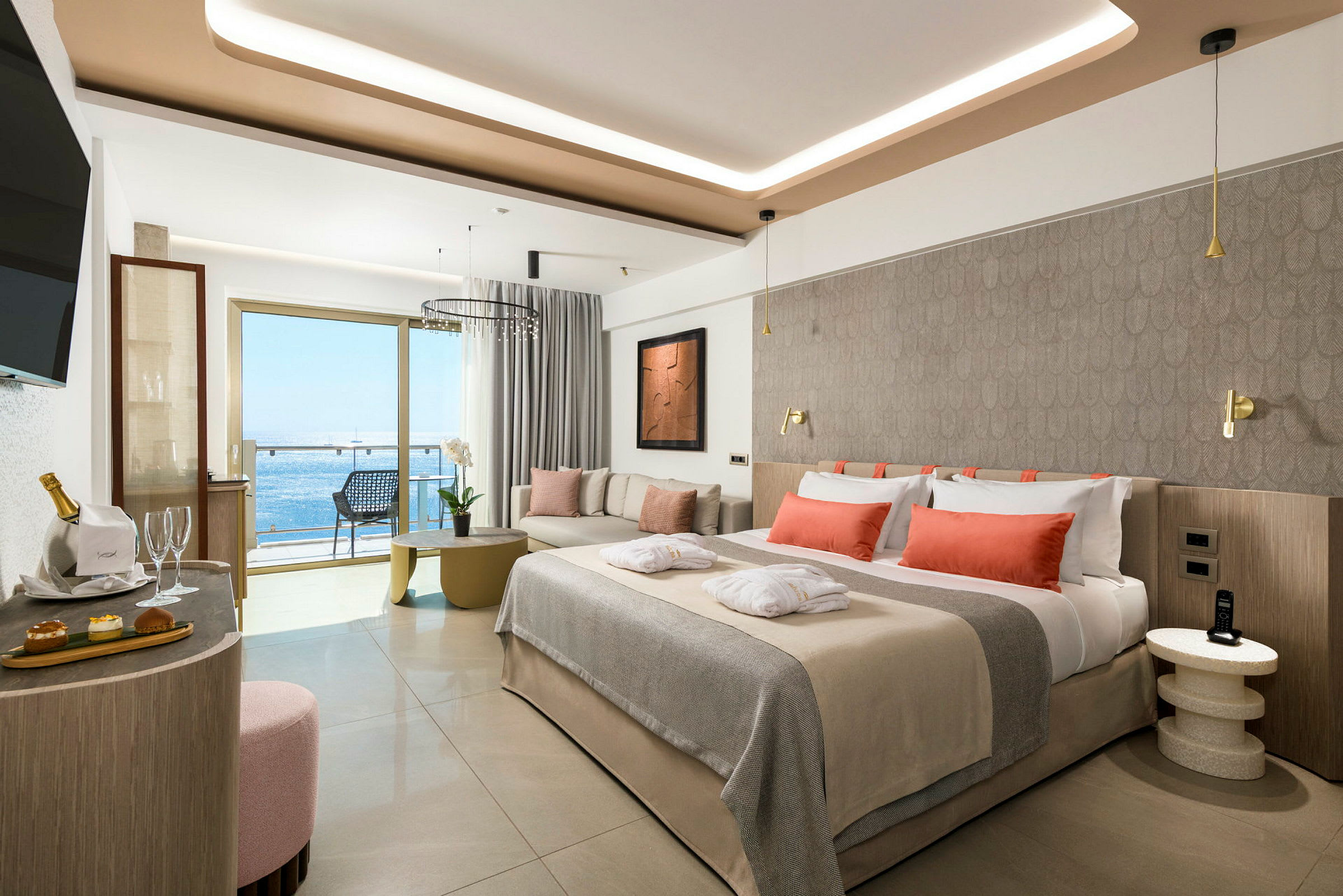 En moderne hotellsuite med oppredd seng med oransje puter, sittegruppe og balkong med havutsikt. (AI-generert tekst)