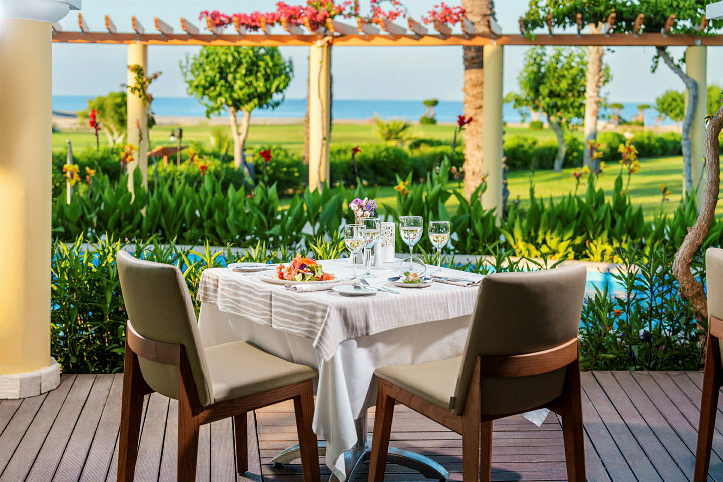 To spisestoler ved et oppdekket bord på en terrasse med utsikt over en hage og havet. (AI-generert tekst)