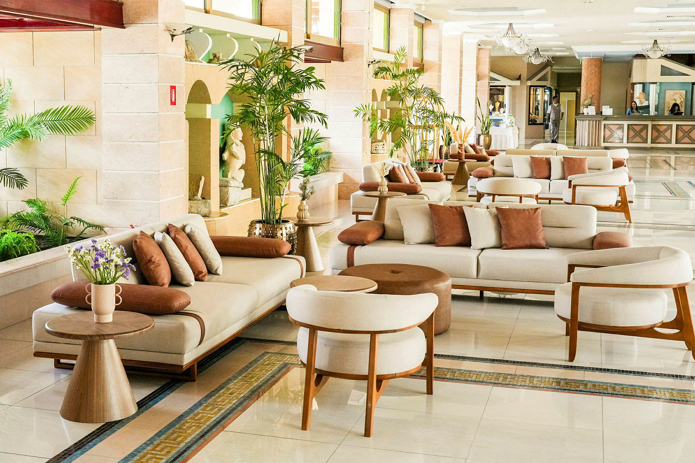 Et hotellobbyområde med hvite sofaer, brun pynteputer og små trebord, omgitt av frodige planter. (AI-generert tekst)