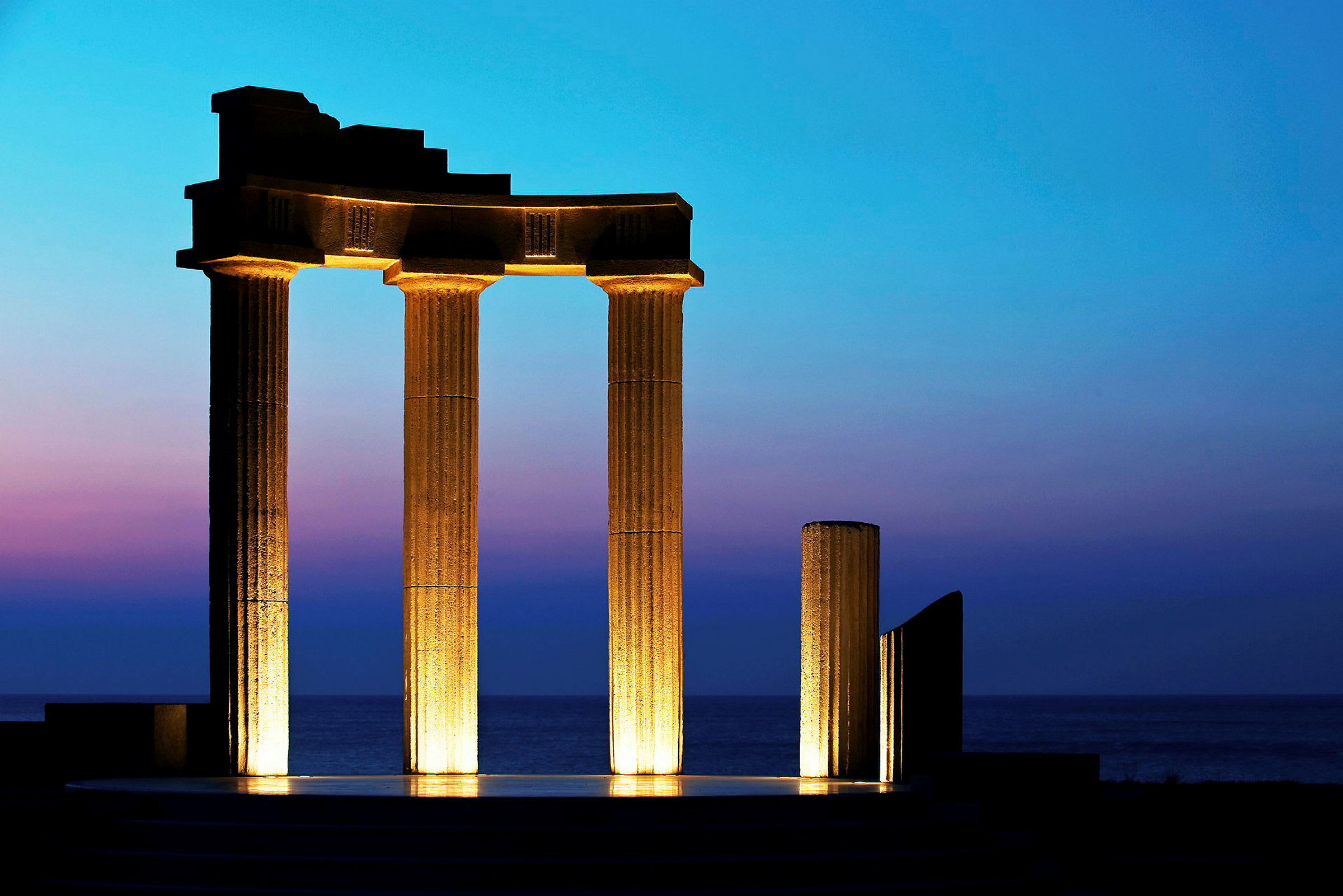 En opplyst antikk søylestruktur foran en blå og lilla skumringshimmel nær havet. (AI-generert tekst)